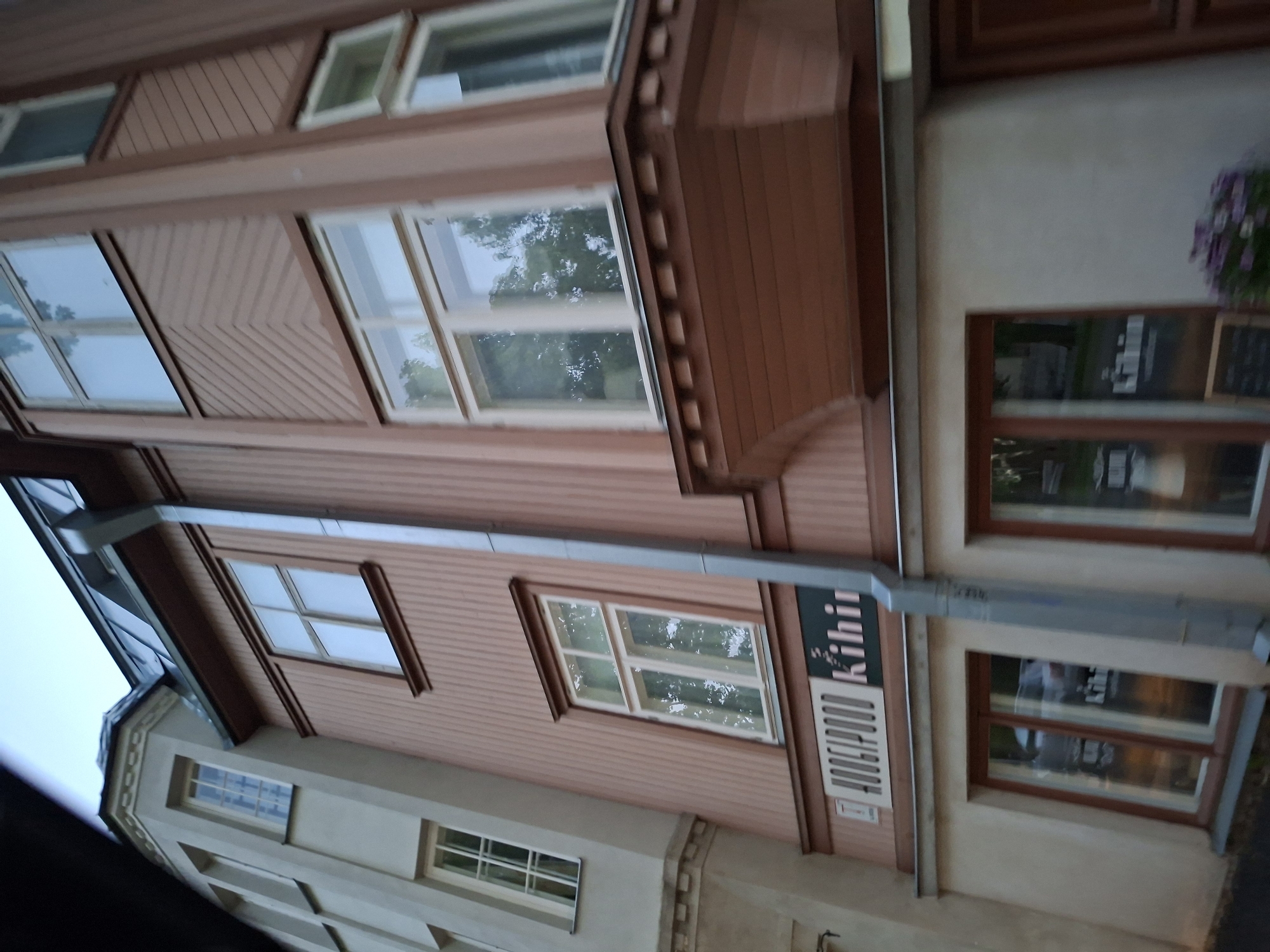 This image depicts a multi-story building in Tallinn, Estonia, specifically identified as Pikk tn 35, home to the "Hoogipood Kihnu" craft store. The building features two distinct architectural sections. The left section, painted in a light beige or off-white, presents a multi-faceted corner leading to a bay window structure on an upper floor, crowned by a dark, possibly metallic, roof. This section includes several vertically oriented, white-framed sash windows.
The right section of the building is characterized by a reddish-brown or terracotta facade with vertical wooden paneling on its upper levels. A prominent darker brown, wooden bay window with decorative cornices protrudes from the upper right, below a series of multi-pane, white-framed windows that reflect trees and an overcast sky. A silver-colored metal downspout runs vertically along this facade. Below the wooden paneling, the ground floor features a lighter, stucco-like finish with large display windows.
A white rectangular sign with "HOOGIPOOD Kihnu" in black lettering is affixed to the reddish-brown facade above the ground floor. Reflections in the ground-floor windows show fragmented text, likely associated with the same shop, as well as a partial reflection of a white vehicle on the street. Near the bottom right, a dark chalkboard-style sign is partially visible in front of a window, alongside a small pot containing purple flowers. The sky is overcast and light grey, indicating diffuse daylight. No individuals are visible.