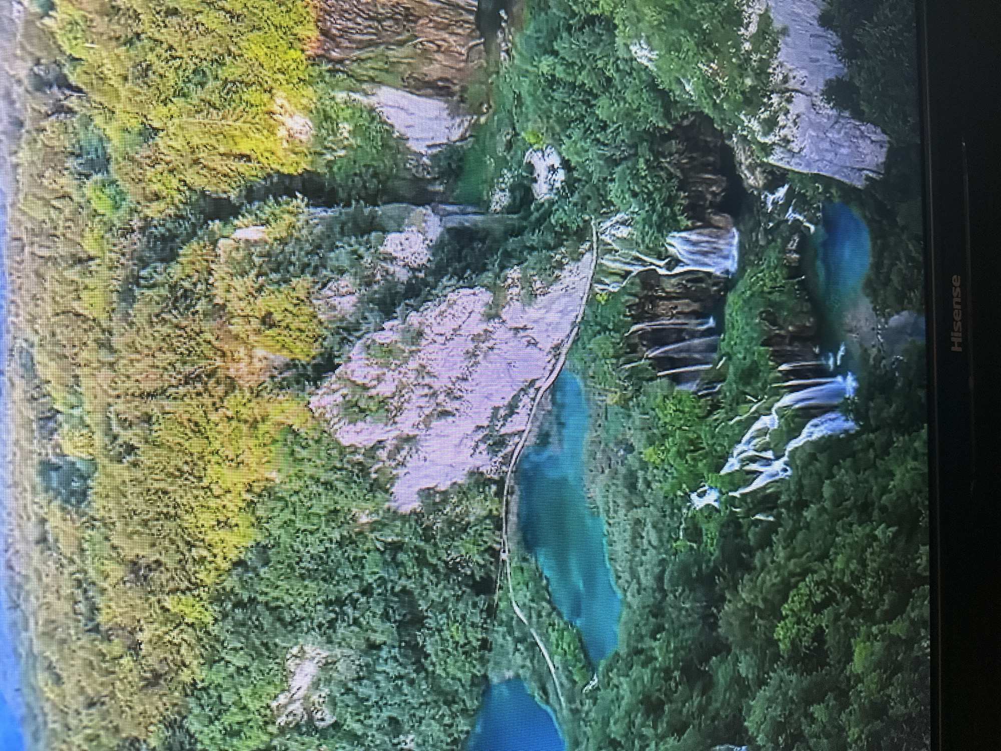 This is an aerial, long shot of a lush, green mountainous landscape with clear turquoise lakes and cascading waterfalls. The image is grainy and appears to be taken from a screen, possibly a television. There are no people visible in the scene. The landscape features dense vegetation covering the slopes and cliffs, with the water bodies nestled within the terrain. The lighting suggests it is daytime, with sunlight highlighting parts of the foliage. The bottom of the image shows a Hisense logo, indicating it is being displayed on a Hisense television. The scene does not depict any elements that would identify it as being located in the city of Kyiv, Ukraine.