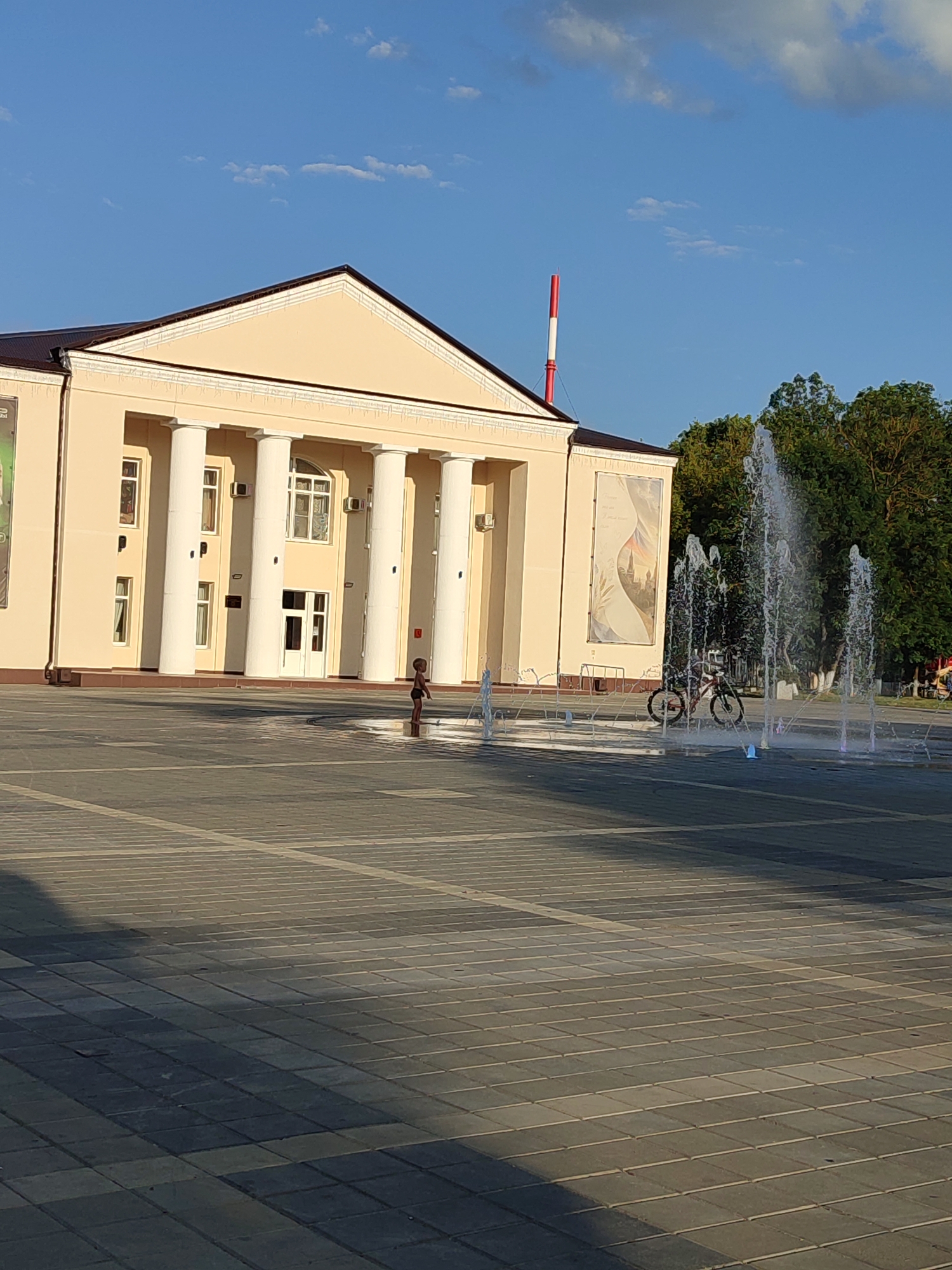 The image shows a wide, paved public square in front of a building with classical architecture, featuring a portico with large white columns. The building is light beige and has a dark-colored roof. A tall, red and white striped smokestack is visible behind the building to the left, and a lush green tree line borders the right side of the scene.
In the foreground, a modern fountain sprays water upwards. A child, dressed in dark shorts, stands within the fountain's spray zone, seemingly interacting with the water. To the right of the fountain, a bicycle is parked. The square itself is paved with large, square tiles, with some tiles appearing wet from the fountain's spray, creating a glistening effect. Sunlight casts distinct shadows across the pavement, indicating a clear, sunny day with the sun positioned at an angle. The sky is blue with scattered white clouds. The building's facade has large windows and multiple entrances. A large, framed artwork is mounted on the exterior wall of the building between two columns. The general atmosphere suggests a public space during daytime, possibly a town square or plaza.