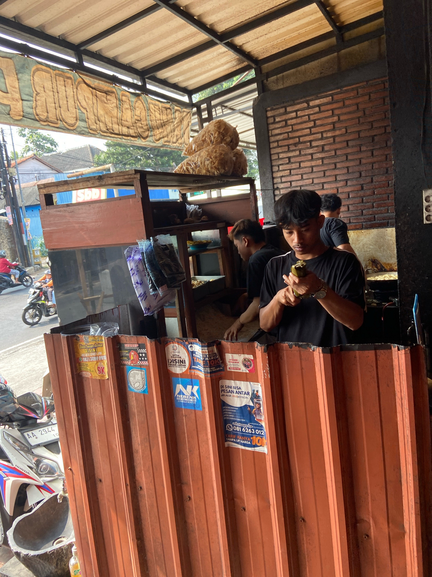 The image depicts a street-side food stall in Salatiga, Indonesia, under a corrugated metal roof supported by black metal beams. A large, faded banner with text is visible above the stall. The stall itself is constructed with wooden planks and has shelves displaying packaged items and what appear to be large piles of fried snacks.
In the foreground, a young man wearing a black t-shirt is standing, holding a piece of food in his hand and examining it. He is wearing a watch on his left wrist. Behind him, another young man in a black t-shirt is partially visible, seemingly working at the stall. Further back, a third person in a black t-shirt is also visible.
The stall is fronted by an orange corrugated metal barrier, plastered with various stickers and flyers. Some of these flyers have text that appears to be in Indonesian, including contact information and promotional messages.
To the left of the stall, a street scene is visible. Two people are riding motorcycles, one in a red jacket and another in a yellow jacket. A white motorcycle with license plate "AA 2944 10" is parked to the left of the stall, partially obscuring a stone-like structure that seems to be a sink or basin with a tap. The environment appears to be urban, with buildings and trees visible in the background. The lighting suggests it is daytime, possibly late afternoon due to the shadows.