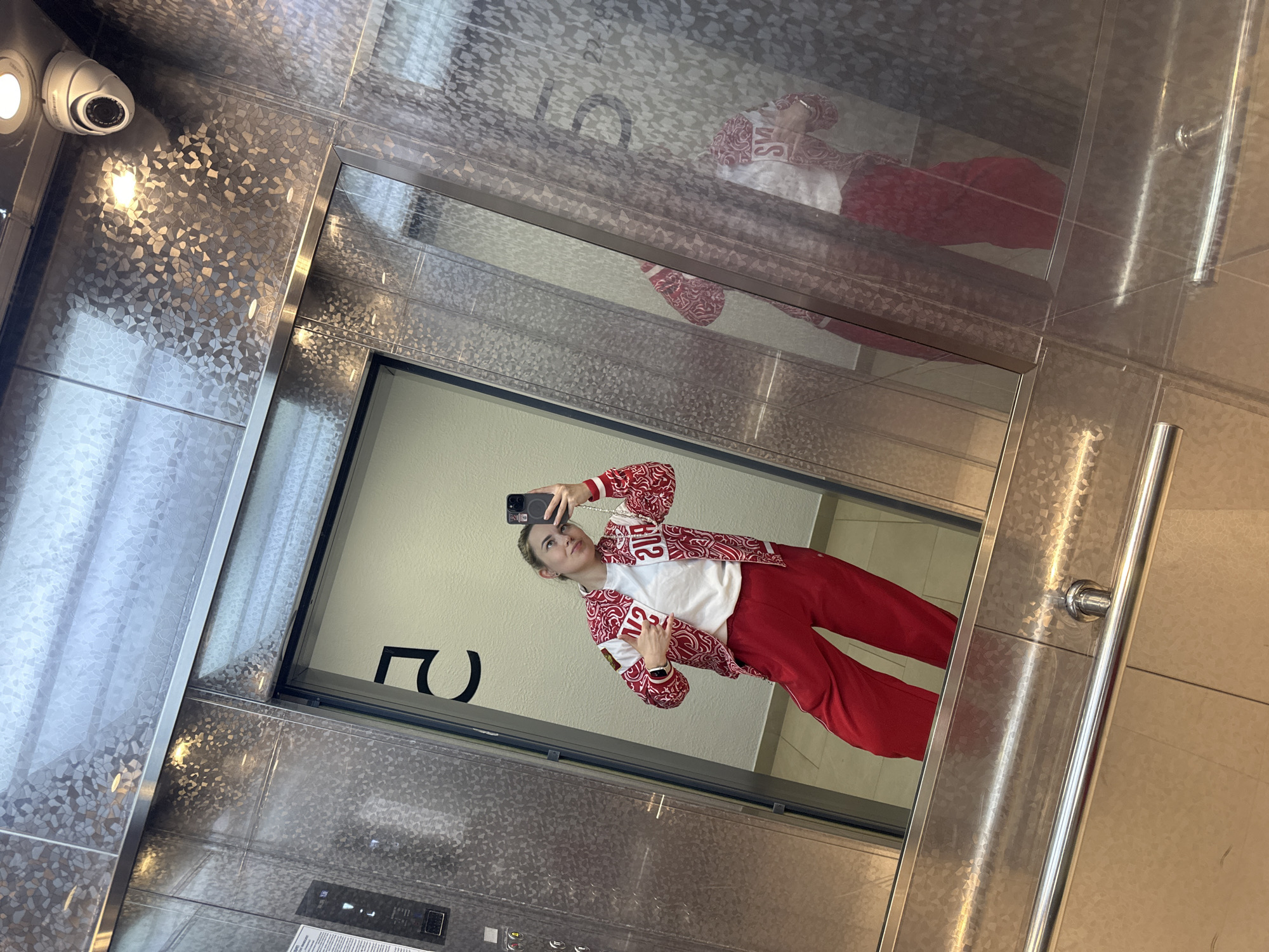 A young woman is captured taking a mirror selfie inside what appears to be a modern elevator or a space with reflective, patterned metallic walls. She stands upright, holding a dark-colored smartphone to photograph her reflection. Her light-colored hair is pulled back, and she has a neutral to slightly contemplative expression, looking towards the phone screen.
She is dressed in a distinctive red and white outfit. She wears a white top underneath a red and white patterned jacket. The jacket prominently features the word "РОССИЯ" (Russia) in Cyrillic lettering on the front, and "РУС" (RUS) on her left sleeve. Her attire is completed with plain red pants.
The setting is characterized by shiny, textured metallic surfaces that reflect the environment. A security camera is mounted on the patterned ceiling in the upper left of the frame. An elevator control panel with illuminated buttons is visible at the bottom, and a metal handrail runs along the right side of the mirror. Behind the woman's reflection, a large black character, identified as the number '2' (likely part of a floor number '22'), is visible on the wall.
The activity is a casual self-portrait session within an indoor public or semi-public space. The lighting is bright and consistent, indicative of artificial illumination. The overall scene suggests a contemporary building interior. The location context for this image is Perm, Russia.