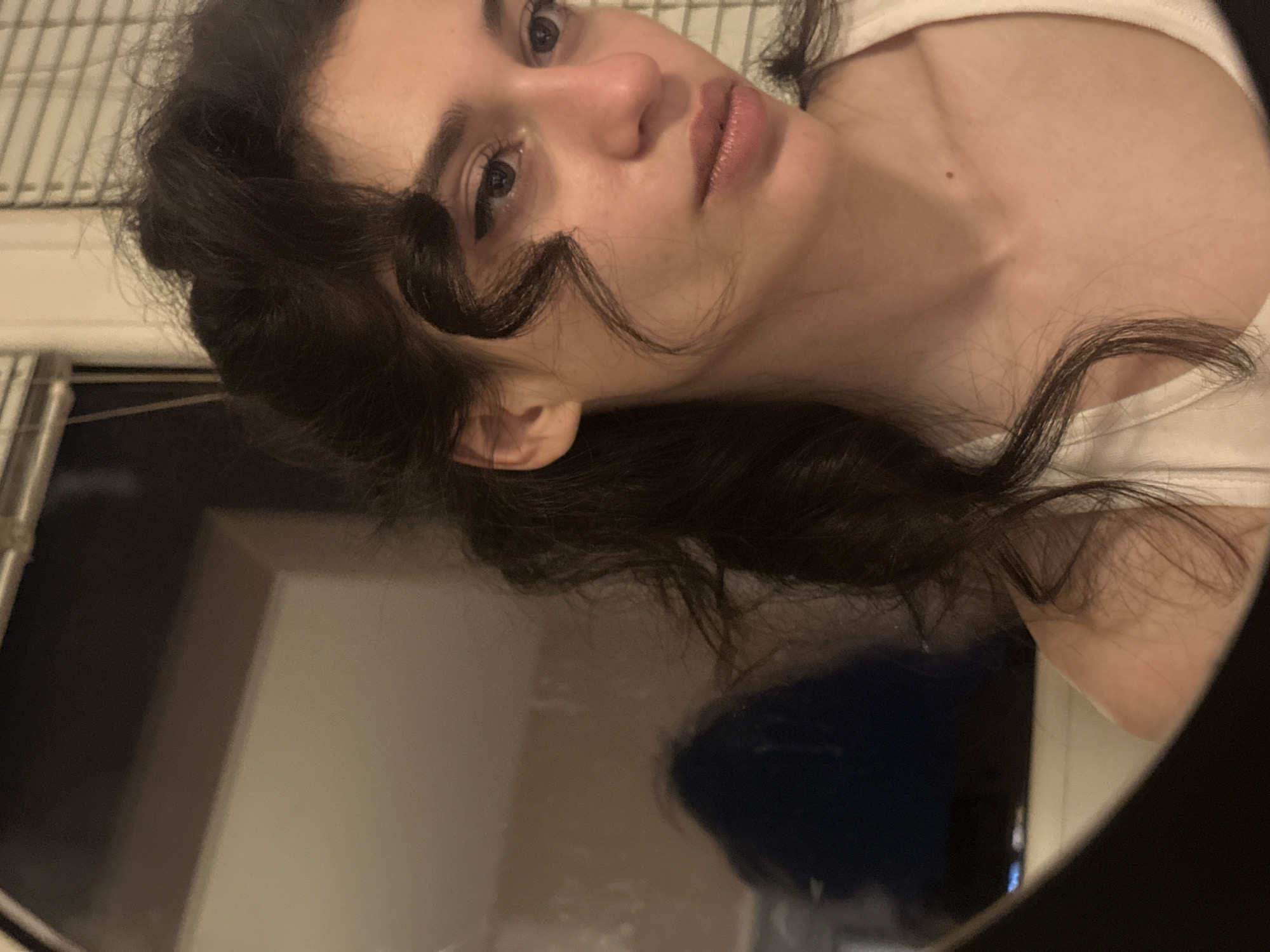 The image shows a young woman with dark, curly hair looking into a mirror. She is wearing a white tank top. Her reflection in the mirror is visible to the left of the frame, showing a dark, blurry figure and what appears to be a window with blinds. The setting appears to be an indoor environment, likely a bathroom or bedroom, given the presence of a mirror and what looks like a wall and blinds. There are no discernible elements within the image that would allow for the identification of a specific location in Sofia, Bulgaria.