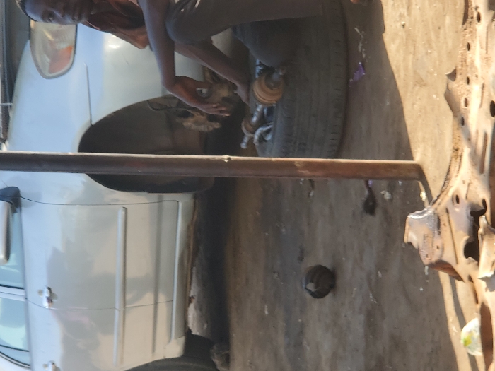 In Kaduna South, Nigeria, a daylight scene depicts an outdoor vehicle repair area. A silver sedan, identified as a Toyota Corolla, occupies the left side of the frame, with its front passenger-side wheel removed, exposing the brake caliper and hub assembly. A young male, wearing a rust-colored shirt and dark trousers, is seated on the unpaved, dusty ground in the mid-right. His right hand rests on his knee. Adjacent to him, a detached black car tire lies flat, supporting several metallic automotive components, including a drive shaft. A dark, vertical metal pole stands centrally. The immediate foreground shows a large, rusted, irregularly shaped metal car part and a dark spherical object on the ground. Scattered debris is present across the dirt surface. Shadows indicate direct sunlight.