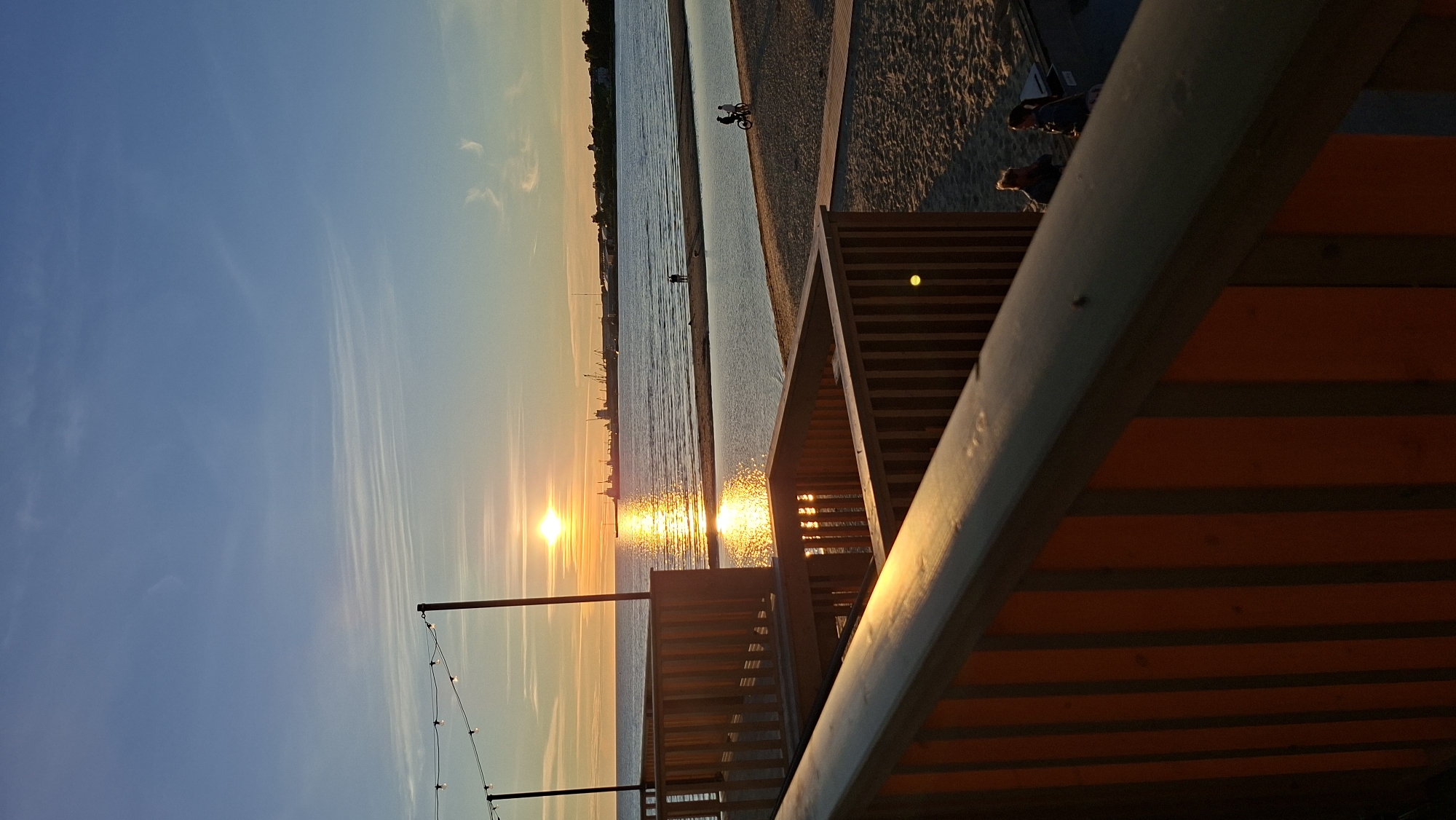 This is a vertical, eye-level shot of a sunset over the sea, taken from a wooden structure at a beach. The sun is a bright orange orb low on the horizon, casting a shimmering golden reflection across the water. In the distance, a coastline with industrial structures and ships is visible. The foreground is dominated by the wooden railings and supports of the elevated structure, with a glimpse of sand and a small body of water. Two people are standing on the beach further away, and two individuals are cycling on a path. A string of lights is suspended above the wooden structure. The sky is a clear blue with wispy white clouds, transitioning to a warm orange near the horizon. The scene appears to be from Pirita beach in Tallinn, Estonia, given the characteristic landscape and presence of industrial elements in the distance, which are common in the Tallinn Bay area.