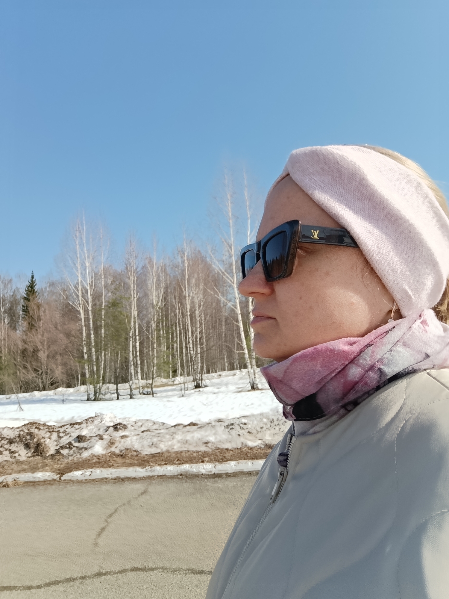 The image depicts a woman in the city of Марковский, Russia, outdoors on a sunny day. She is wearing a light pink headband, large dark-colored sunglasses with a "LV" logo on the temples, and a light-colored jacket. A patterned pink and white scarf is visible around her neck, underneath the jacket.
The background includes a snow-covered area with several bare trees. The sky is a clear blue. A portion of a paved road is visible in the foreground.