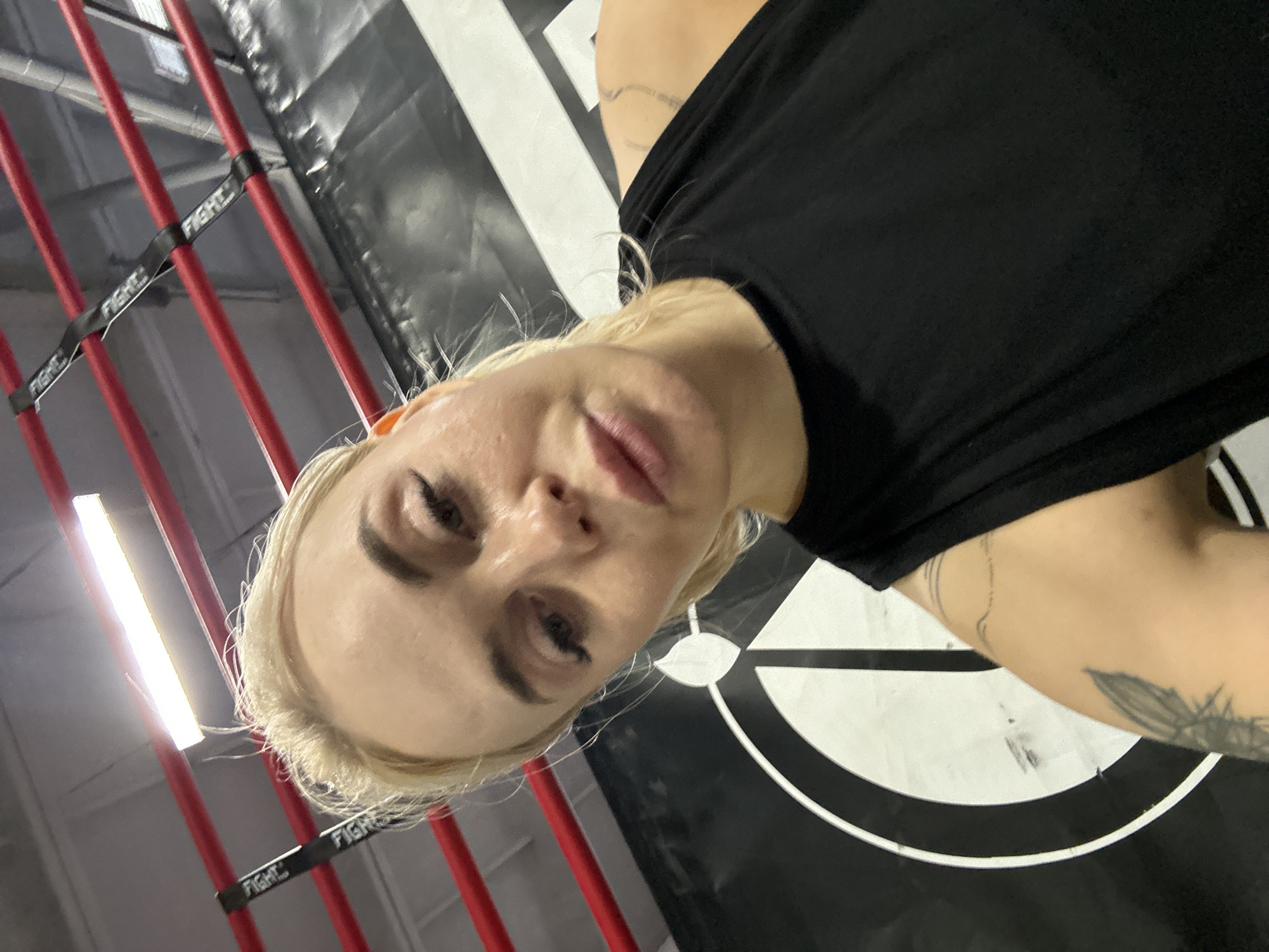 A woman with blonde hair, wearing a black top, is standing inside a fighting ring. The ring is composed of red vertical bars connected by black straps marked "FIGHT". A black and white target is visible in the background. The image appears to have been taken indoors, possibly in a gym or training facility located in Megion, Russia.