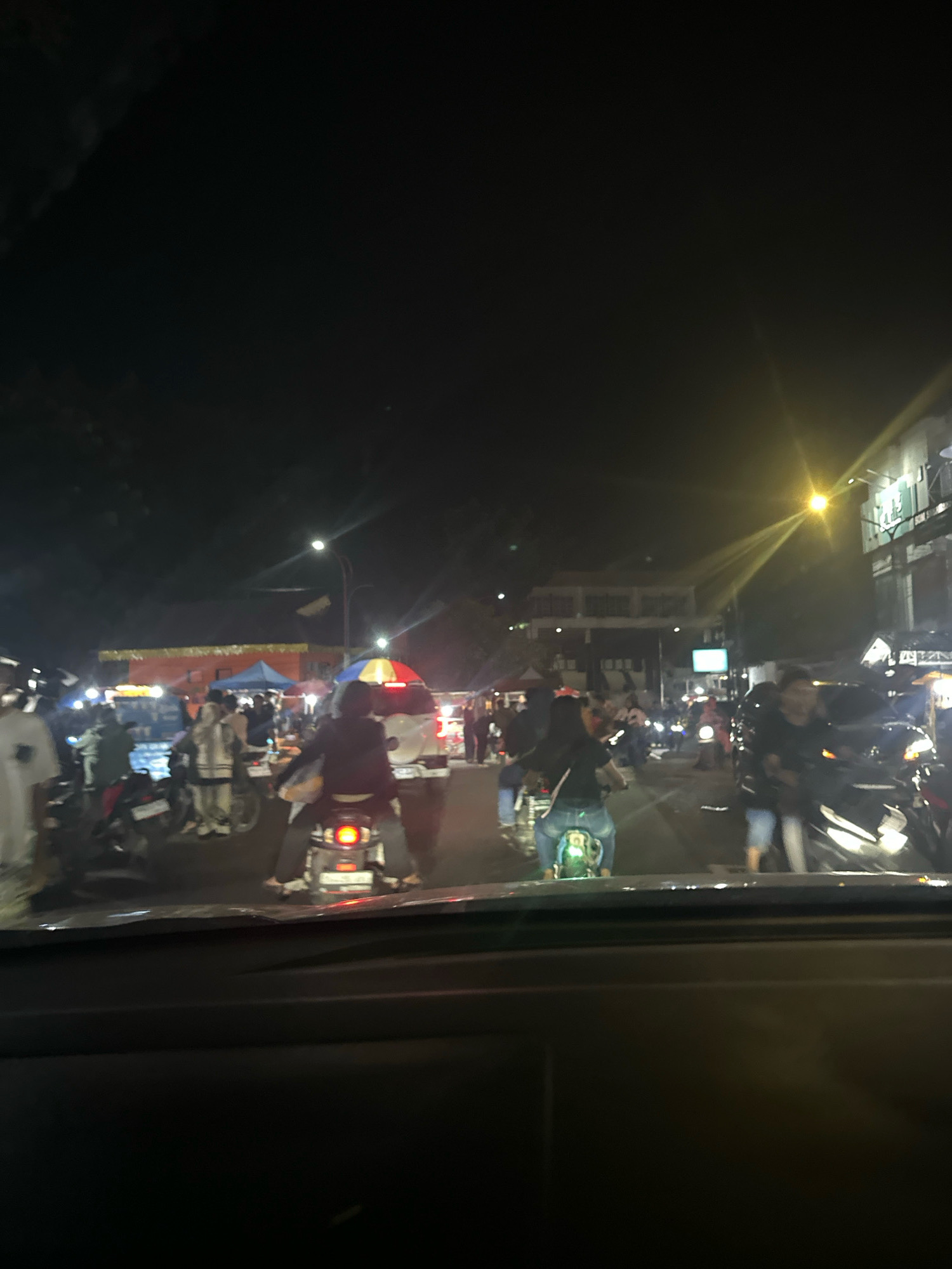 A night-time outdoor scene captures a bustling street scene in the city of Binjai, Indonesia. The foreground is dominated by the dark interior of a vehicle, likely a car, with its dashboard visible. Beyond the vehicle's windshield, a busy street unfolds, illuminated by streetlights and the lights of various stalls and buildings. Numerous people are present, many of whom are riding motorcycles, indicating a common mode of transportation in the area. Some individuals are walking, and stalls suggest a marketplace or event is taking place. The atmosphere appears lively and active, typical of a night market or a busy urban street at night. The architecture visible in the background, though somewhat obscured by darkness, includes buildings that appear to be shops or establishments. The presence of a brightly colored umbrella over a stall adds a touch of visual interest to the scene.