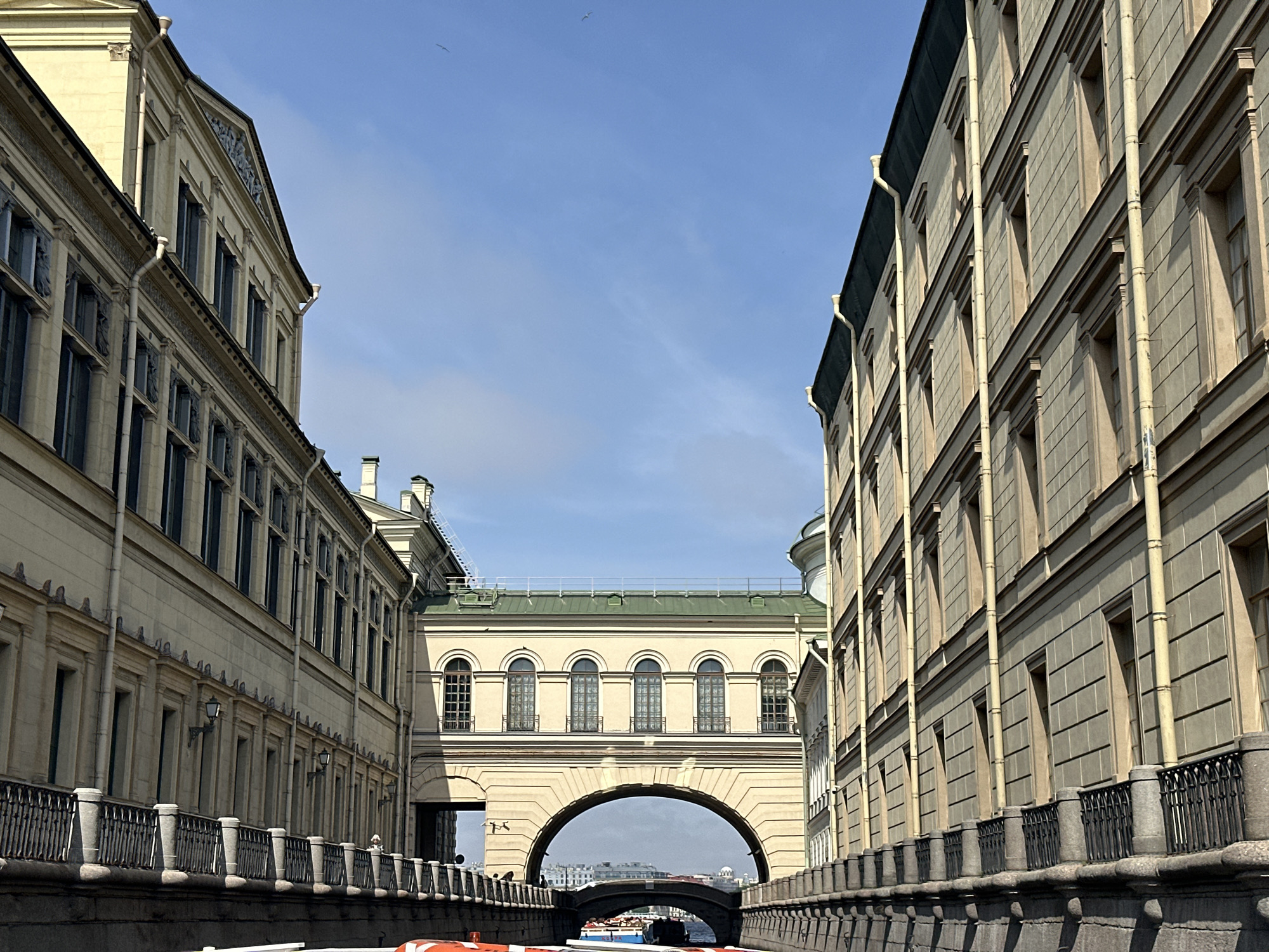This image shows a canal in Saint Petersburg, Russia, framed by buildings on either side. A bridge with an archway spans the canal. Above the archway, a covered walkway connects the two buildings. Several birds are flying in the sky. A boat is visible in the canal, passing beneath the bridge. The buildings are ornate and classical in style, constructed from light-colored stone. Iron railings line the canal banks.