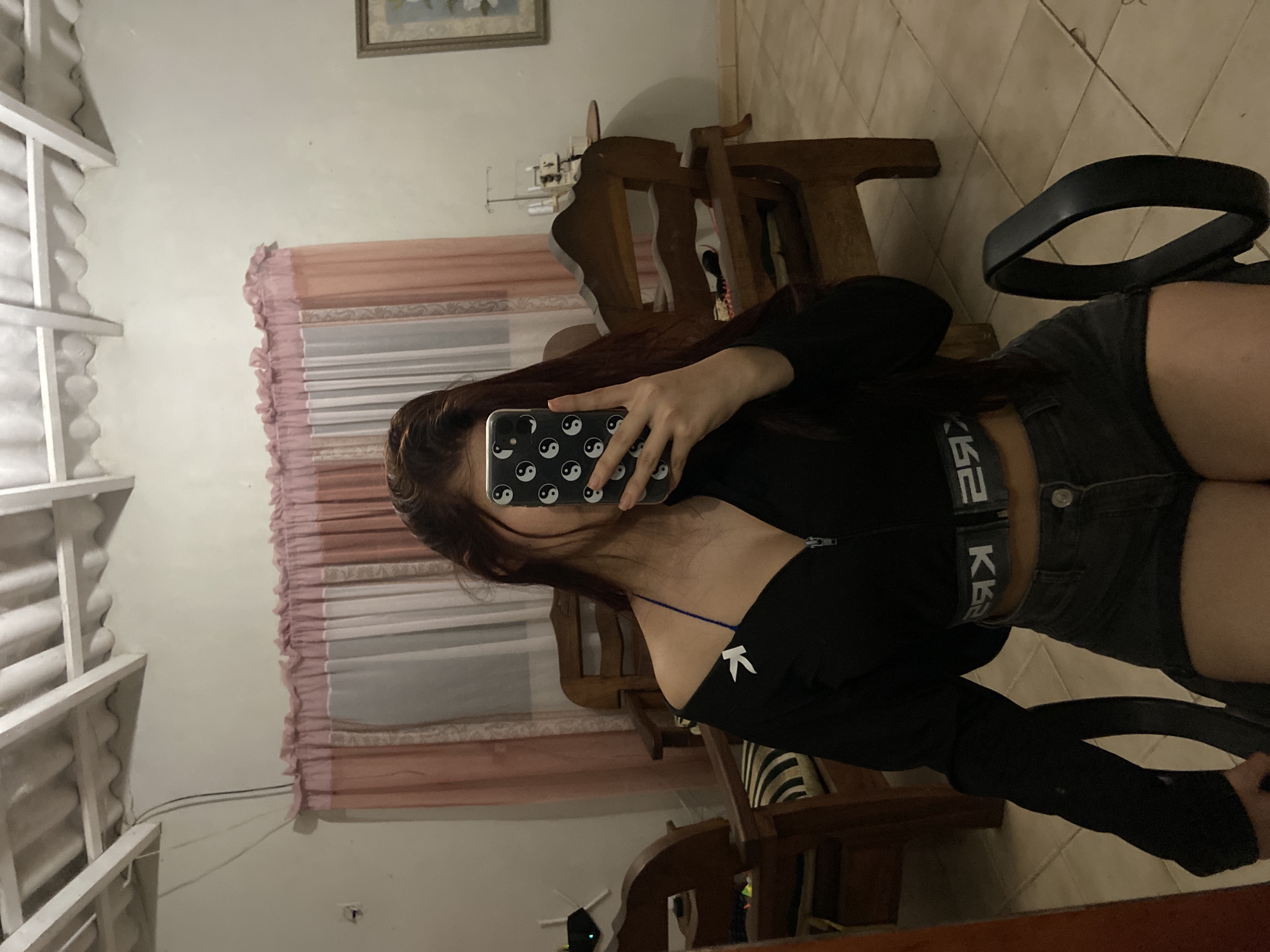 A young woman is taking a mirror selfie. She is sitting in a black chair, wearing dark denim shorts and a long-sleeved black crop top. She is holding a smartphone with a black and white yin-yang patterned case in front of her face. The environment includes a room with a wooden chair visible behind her and a window with blinds. The room appears to be in a home. No information indicates the image was taken in Juan Griego, Venezuela.