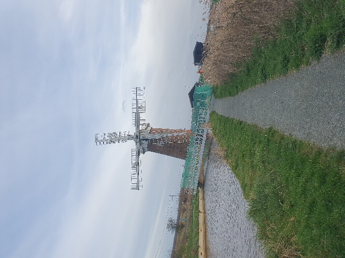 The image depicts a static rural scene featuring St Benet's Abbey Windpump near Ludham, North Norfolk, United Kingdom, under an overcast sky. A gravel path bisects the foreground, bordered by short green grass on the left and taller, dry reeds on the right. To the left of the path, a calm body of water, consistent with a drainage canal, flows. A light-colored metal bridge, partially obscured by green netting, spans this waterway, leading towards the base of a prominent brick tower windmill in the mid-ground. The windmill possesses distinctive skeletal metal sails. Behind the windmill, to the right, are several dark, low-lying structures. The sky is uniformly grey with diffused cloud cover. No individuals or active subjects are present.