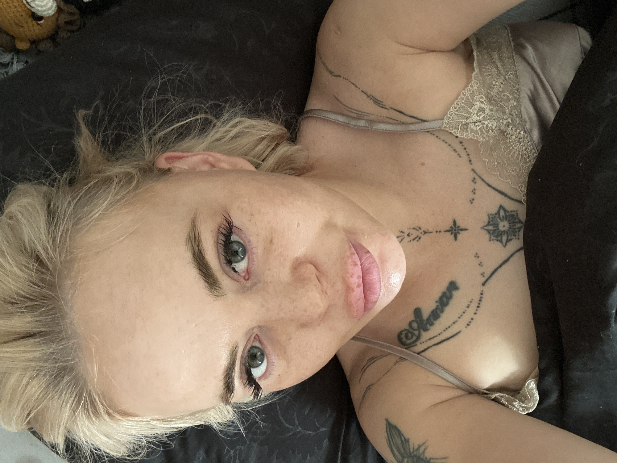 A blonde individual, appearing female, with fair skin and numerous freckles, lies in dark bedding, capturing a selfie from above. Her blonde hair is styled loosely, revealing a clear forehead. She has prominent eyebrows and long eyelashes, complementing her blue-green eyes that gaze directly at the camera. Her full lips are slightly parted in a pout. Visible tattoos include a partial winged design on her left upper arm, script lettering (possibly "Amirian") on her left chest with dotted lines, a central geometric star-like pattern on her chest, and a series of thin, curved lines on her right shoulder. She wears a light-colored camisole with lace trim. In the blurry background on the right, a small, dark-brown crocheted animal head with a white snout is partially visible against the dark fabric.