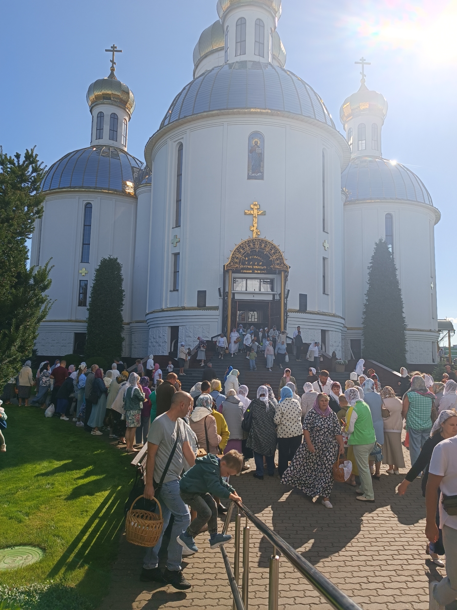 The photograph captures a bright, sunny day in front of a large, white Orthodox church with golden domes, located in the city of Brest, Belarus. A crowd of people, predominantly women wearing headscarves, are gathered outside the church, many in a queue leading up the steps to the entrance. Some individuals are carrying baskets. The church facade is ornate, featuring golden crosses and decorative elements. The ground is paved with cobblestones, and a green lawn is visible to the left, casting long shadows from trees. The sky is clear blue with a noticeable sun glare in the upper right corner. The overall atmosphere suggests a religious gathering or service.