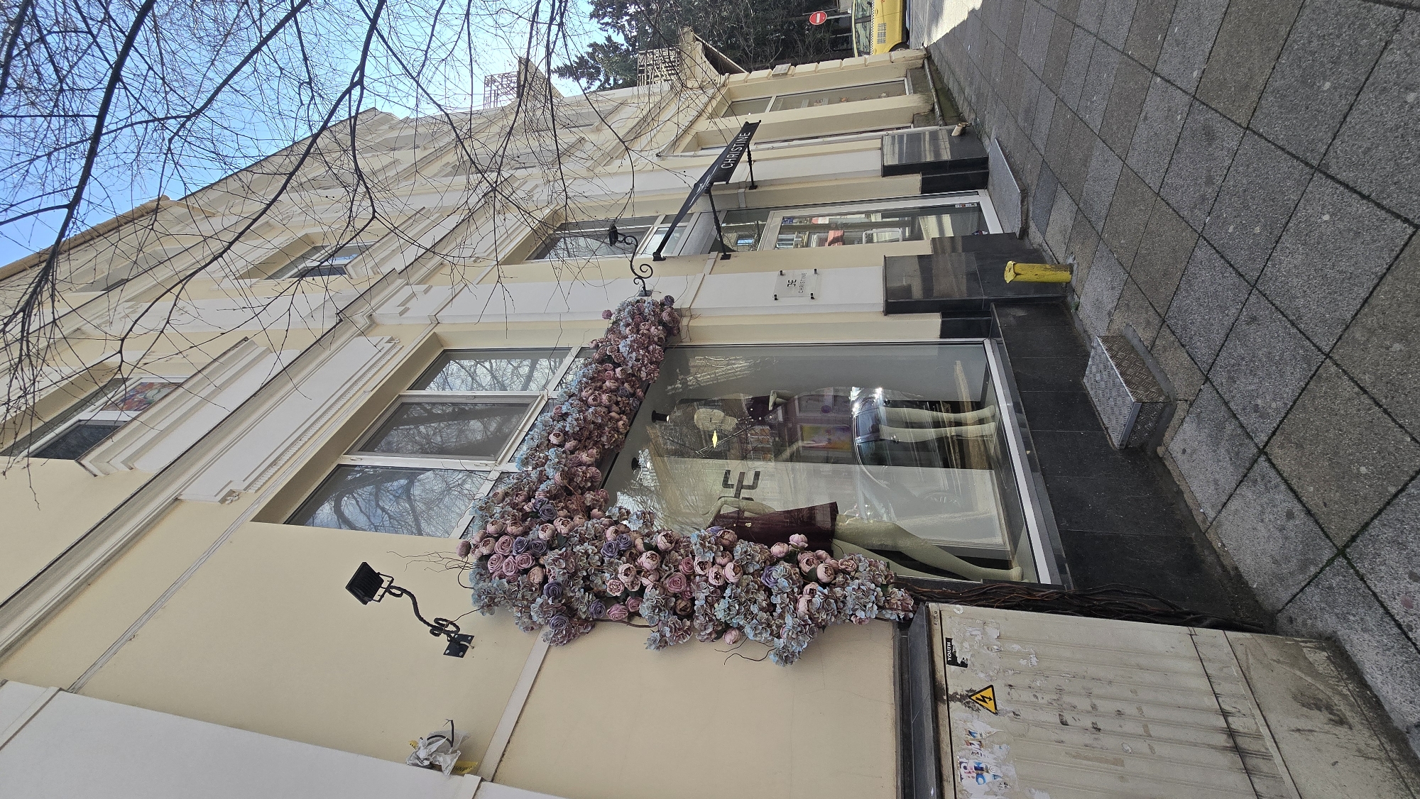 The image shows the exterior of a building in Stara Zagora, Bulgaria. The building has a light yellow facade and multiple windows. A floral arrangement of pink and blue flowers decorates the side of the building adjacent to a large shop window displaying clothing. A black sign with the name "CHRISTINE" is attached above the window. A security camera is mounted on the wall to the left of the floral arrangement. The sidewalk is paved with gray tiles. In the background, there is a glimpse of trees and a yellow vehicle. An electrical box with warning symbols is situated near the bottom right corner of the image.