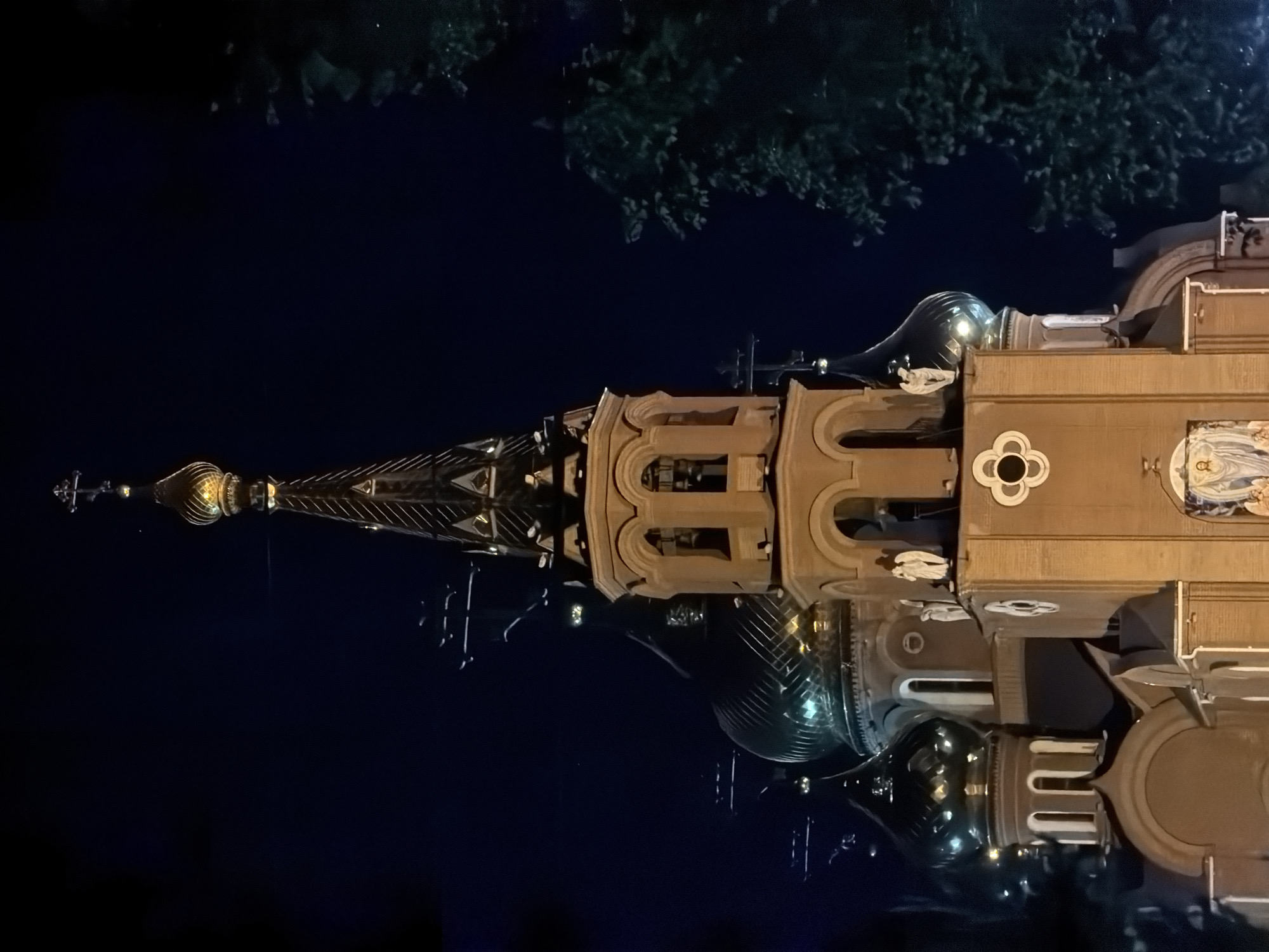 The image shows a building, possibly a church or cathedral, at night. The structure is brightly lit and has a prominent spire with a golden dome at the top. The architectural style is ornate, with several domes and detailed facades. A section of trees appears in the top portion of the image. Based on the architectural style and provided location (Chaikovsky, Russia), it is possible the building is part of a Russian Orthodox church complex. The image is dark and the details in the background are obscured by the night.