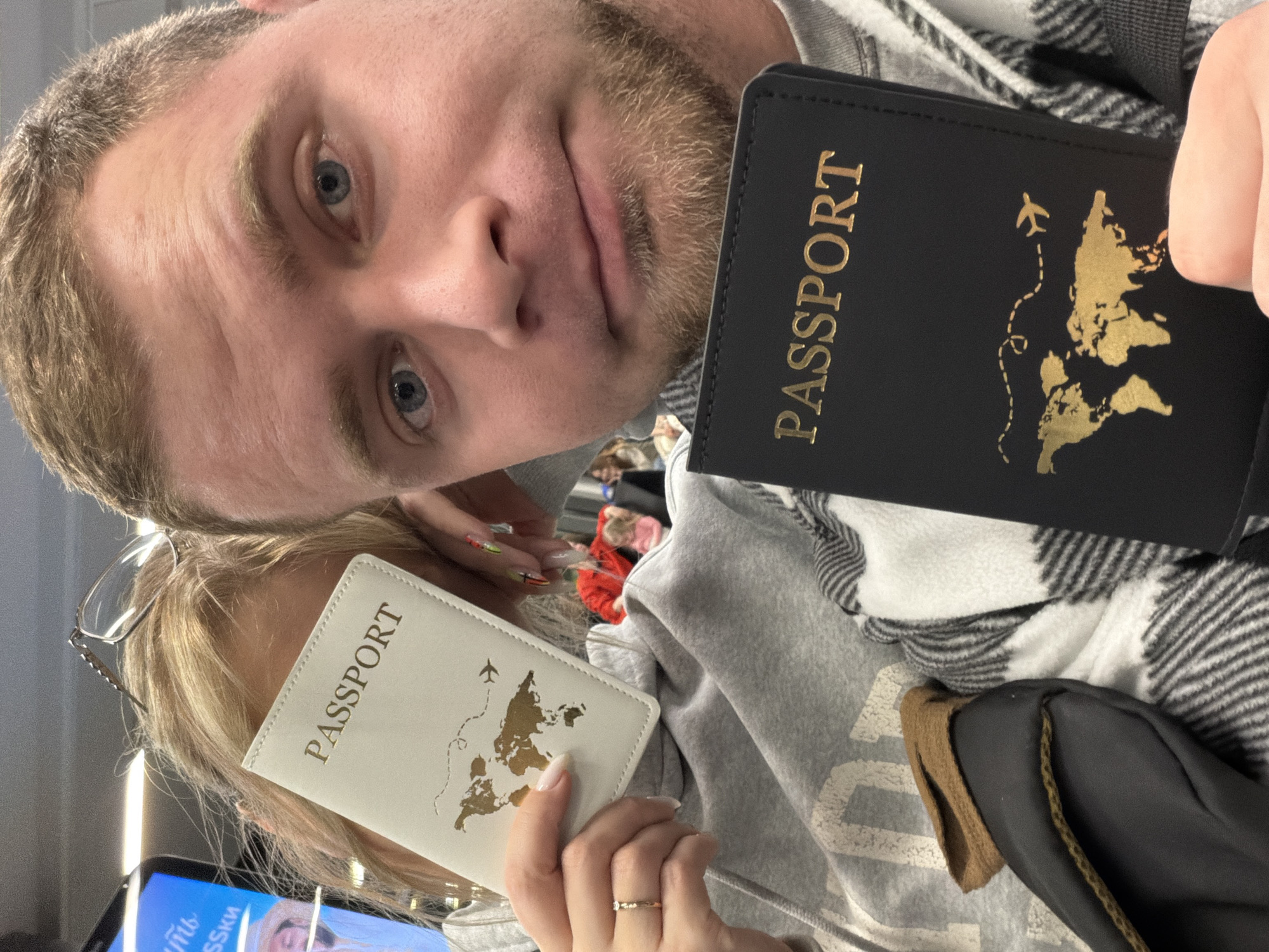The image depicts two individuals in a selfie-style photograph taken within an indoor public waiting area in Zarechnaya (Gamovskoe S/P), Russia. The male subject occupies the right foreground, exhibiting light brown hair, blue eyes, and a light beard, looking directly into the camera. He holds a black passport holder emblazoned with "PASSPORT," a gold world map, and a gold airplane icon. He is wearing a grey top and a black and white plaid garment. To his left, partially obscured, a female subject presents a white passport holder with an identical gold design, concealing her facial features. She has blond hair, glasses positioned on top of her head, and is wearing a grey hooded sweatshirt. A ring is visible on her left ring finger, and her fingernails display a multicolored pattern. The background is blurred, showing additional individuals, one in a red top and another in pink, consistent with a public seating area, along with overhead artificial lighting and a digital display screen containing partial Russian text.