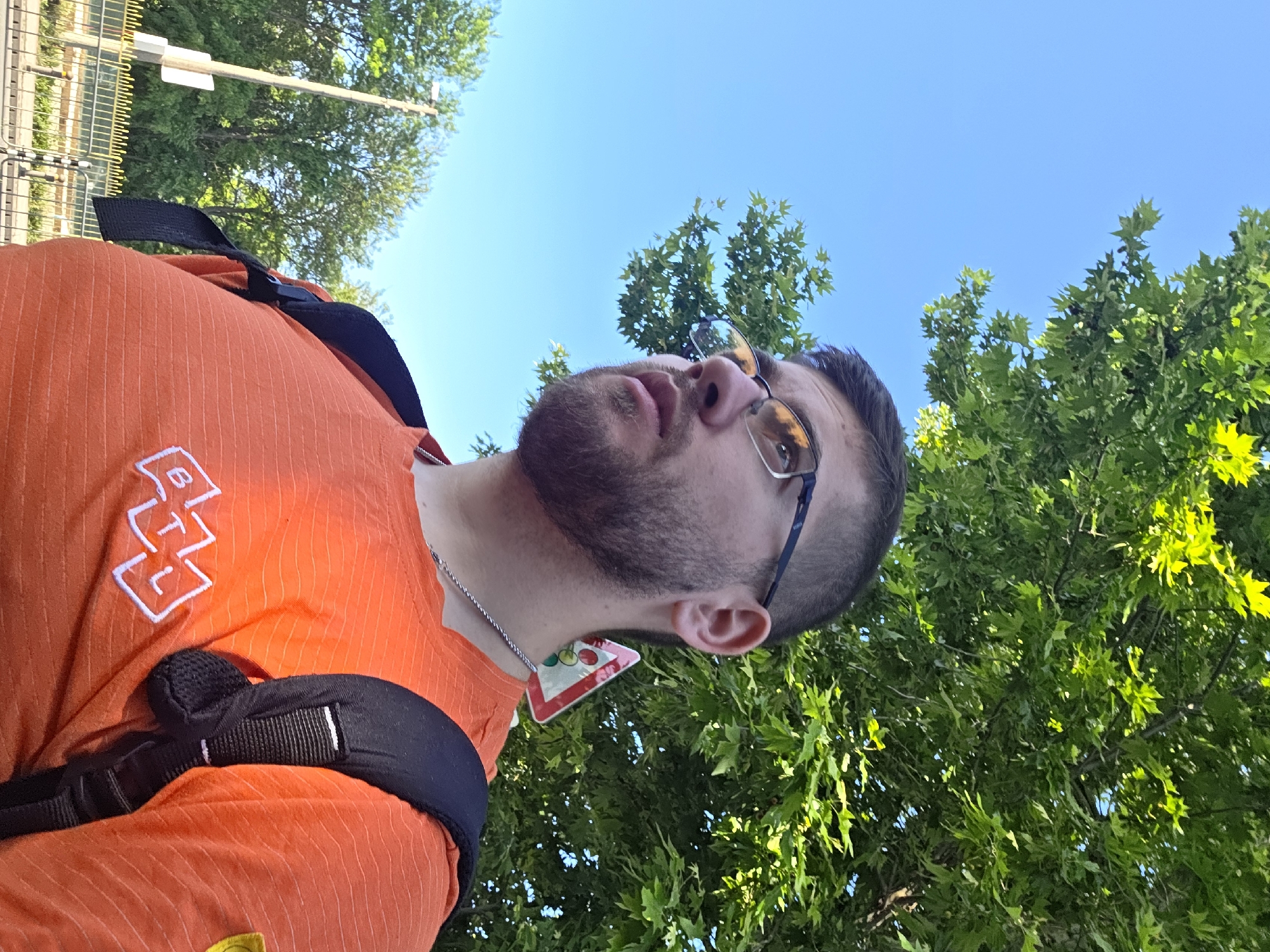 The image shows a man wearing glasses and an orange shirt with a backpack. He is looking up at the sky. In the background, there are trees and a blue sky. The location appears to be outdoors, possibly in a park or near trees in Plovdiv, Bulgaria, although there are no immediate landmarks or details to verify the precise location. The presence of the trees and the lighting suggest it's daytime and likely sunny. A street sign is visible in the background on the left. The man is wearing a silver chain around his neck.
