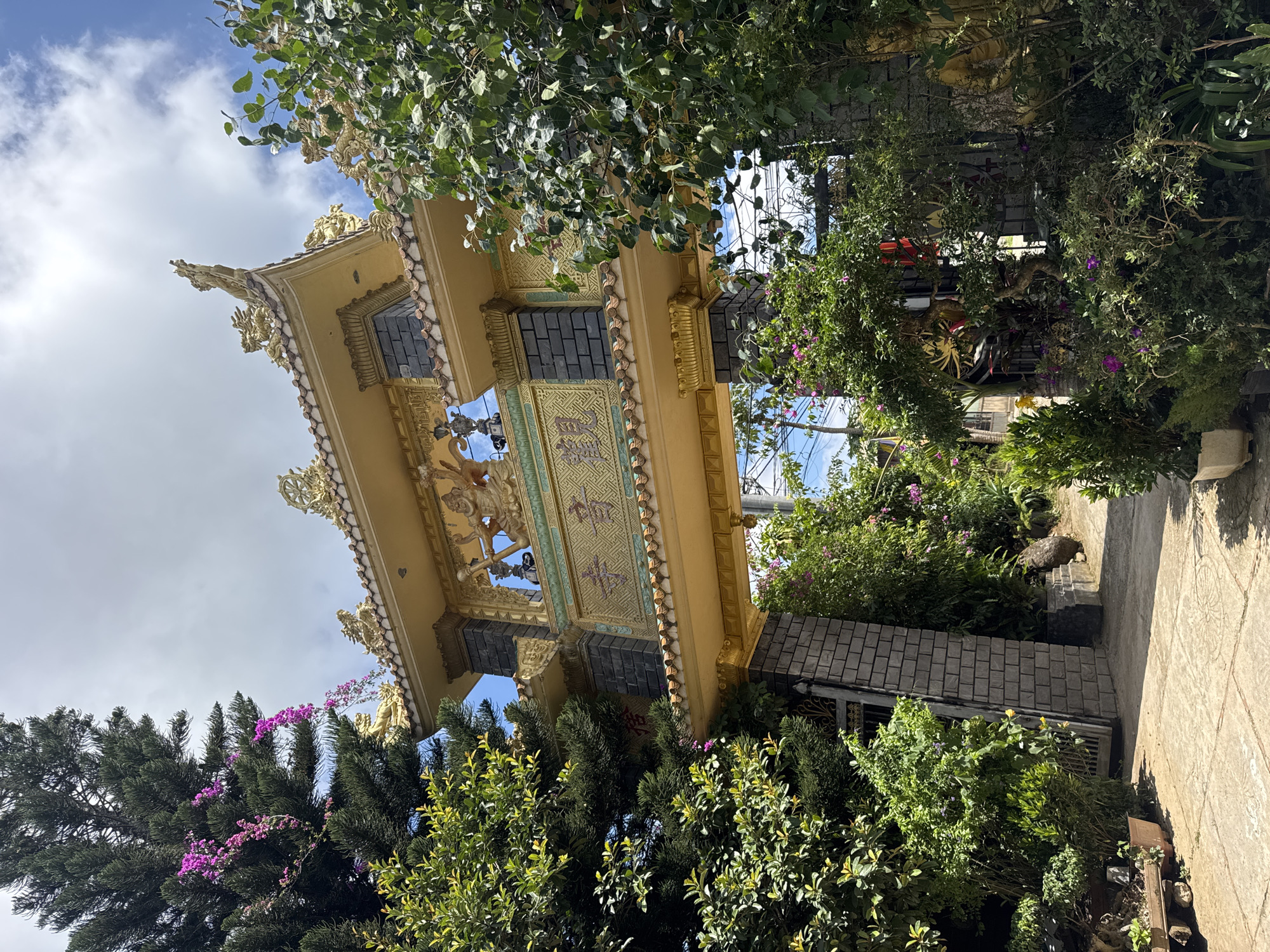 This is a photograph of the entrance gate to a Buddhist temple in Da Lat, Vietnam. The gate is ornate and features gold-colored dragons and other traditional motifs. It is flanked by lush green trees and flowering plants, with a clear blue sky and white clouds overhead. The gate itself is made of stone and brick, with a tiled roof. The overall scene conveys a sense of tranquility and spirituality.