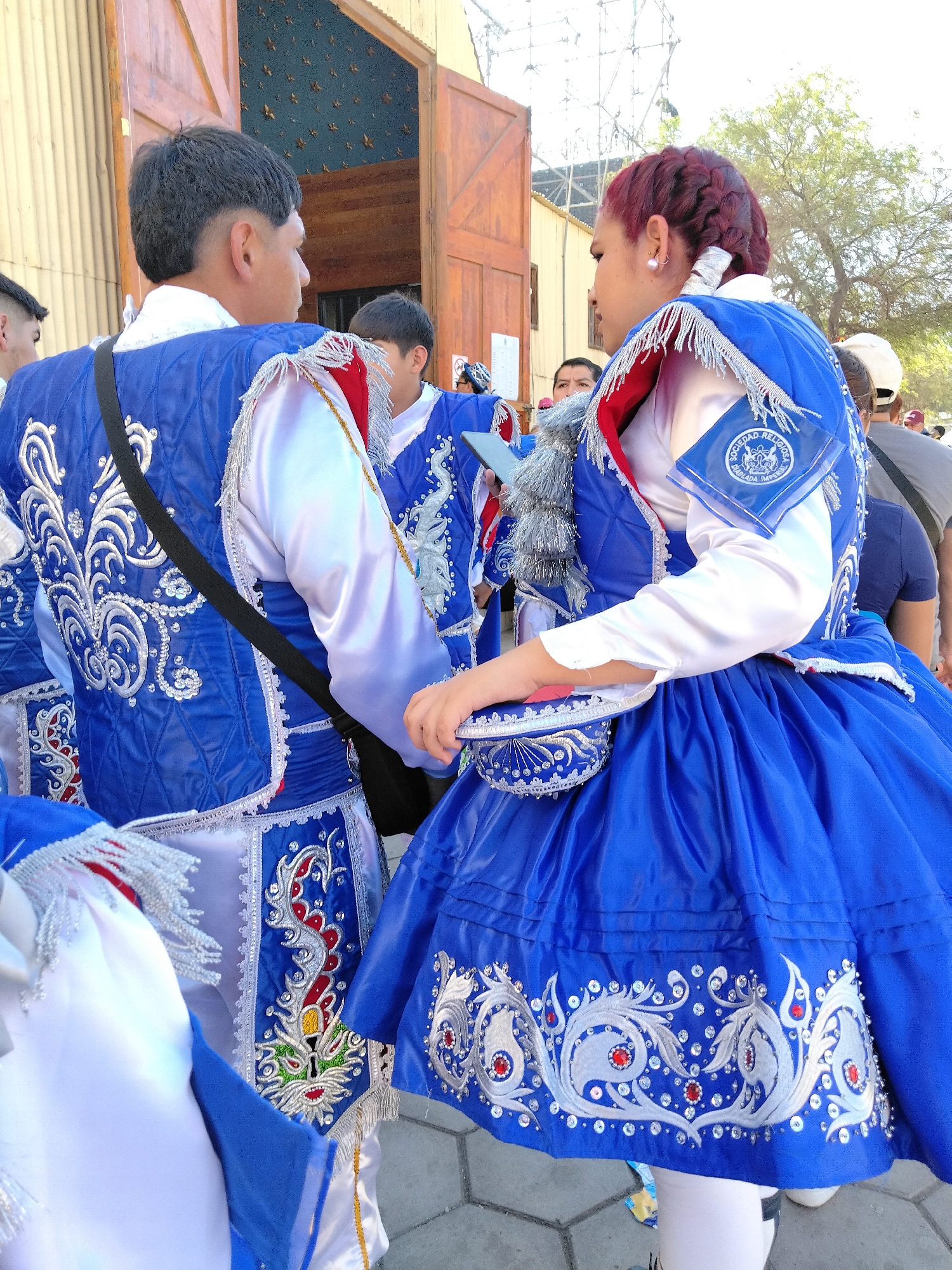 In Pozo Almonte, Chile, a vibrant cultural event unfolds. The foreground features individuals adorned in elaborate traditional costumes, predominantly in rich royal blue, accented with intricate silver embroidery and red detailing. The attire includes ornate vests, flowing skirts, and white sleeves, suggesting a dance or ceremonial performance. One individual's vest displays a patch with the text "SOCIEDAD RELIGIOSA DIABLADA IMPERIAL." Behind them, an imposing wooden doorway with star-like embellishments on its facade stands open, leading into a dimly lit interior. The background reveals a street scene with more people, trees, and scaffolding, indicating an outdoor celebration or gathering in the city. The overall atmosphere is festive and culturally significant, capturing a moment of community and tradition.