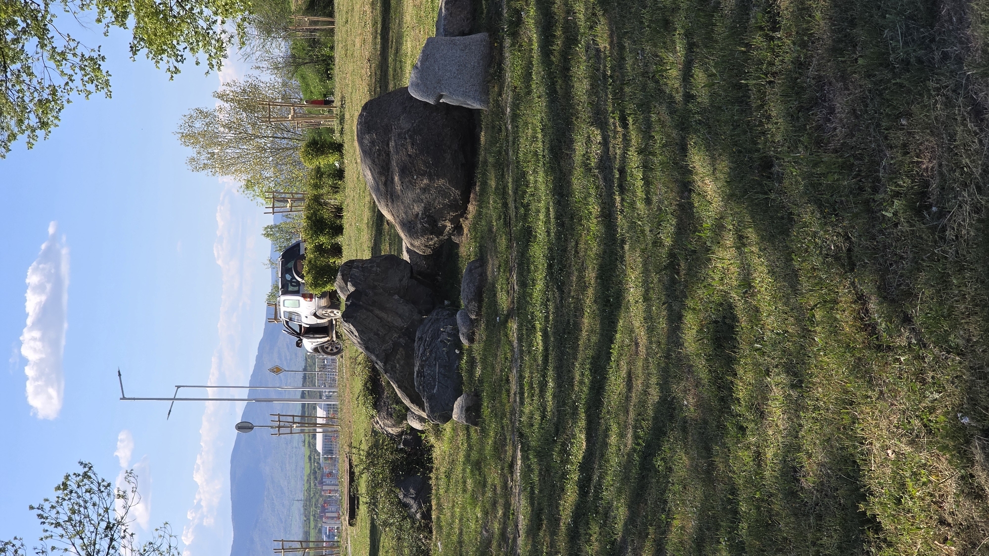 The image shows an outdoor scene in Blagoevgrad, Bulgaria. A white vehicle is parked near a cluster of large rocks on a grassy area. In the background, there are mountains, trees, street lights, and a partial view of the city. The sky is blue with scattered clouds. The grass has shadows.