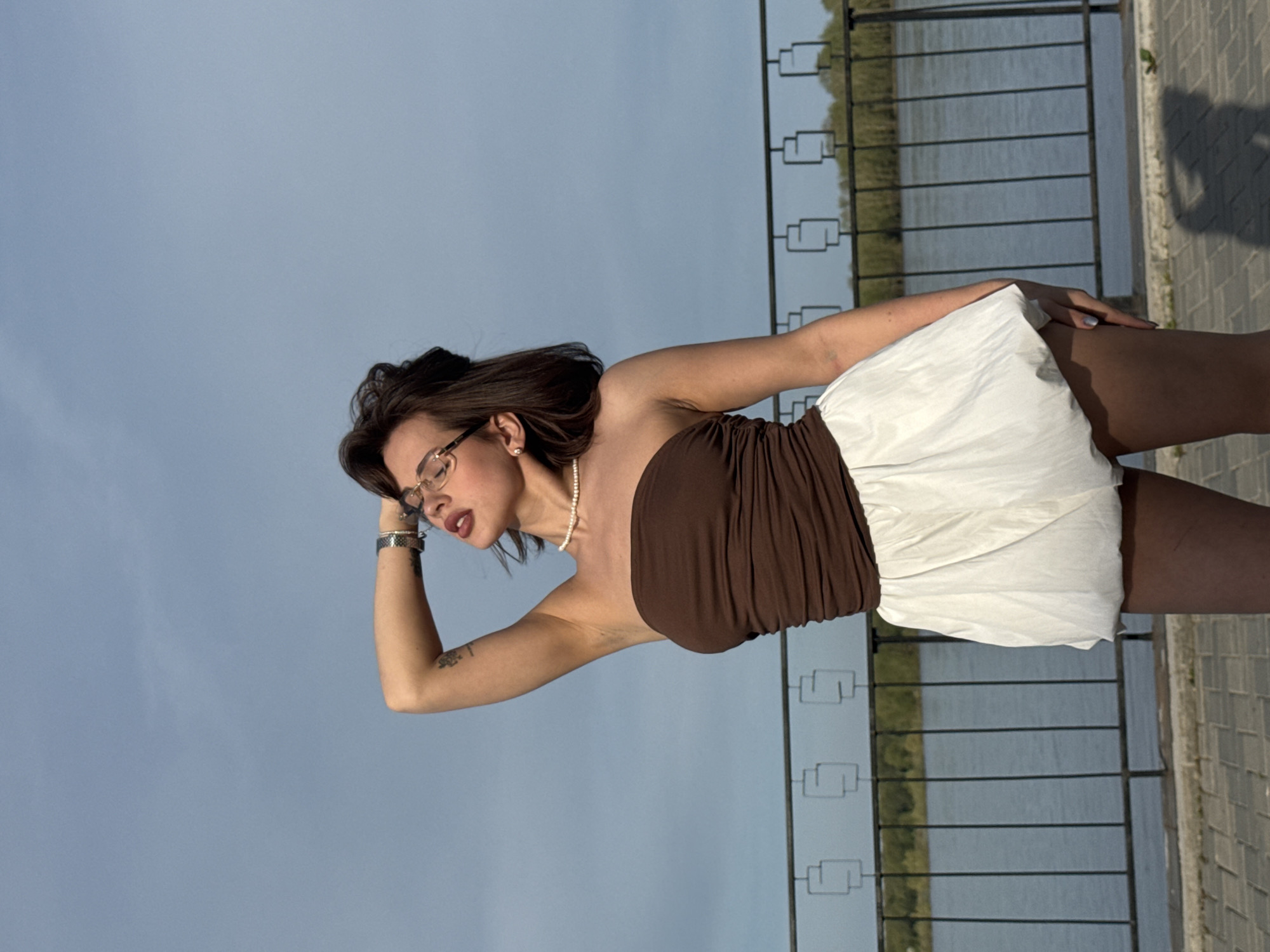 A woman is standing next to a fence at a waterfront location, possibly in Sarapul, Russia. She wears glasses, a brown top, and a white skirt. She is looking away from the camera and has one hand on her head. A body of water is visible beyond the fence. A paved walkway is alongside the fence. The sky is light blue.