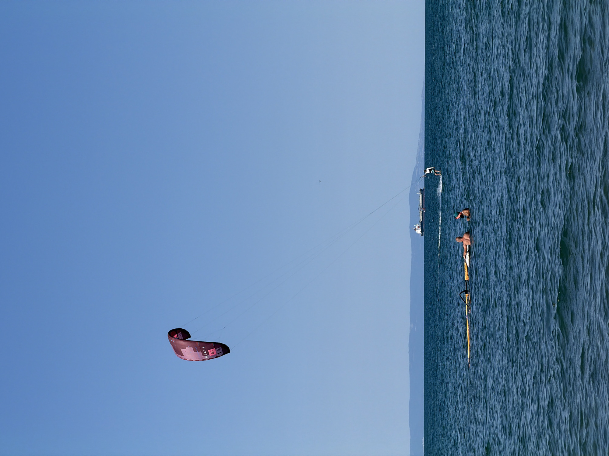 A wide shot captures a bright, sunny day at the coast of Atiya, Bulgaria, with a clear blue sky and calm, deep blue water. In the distance, a faint, hazy mountain range forms the horizon line. On the water, a lone kitesurfer glides across the surface, their kite a vibrant splash of red against the sky. Further out, a large cargo ship and a smaller vessel navigate the sea. In the foreground, two people are partially submerged in the water, one appearing to be resting on a yellow inflatable mat, while another person, wearing a green cap, looks towards the shore. The overall scene conveys a sense of leisure and outdoor activity on a tranquil sea.