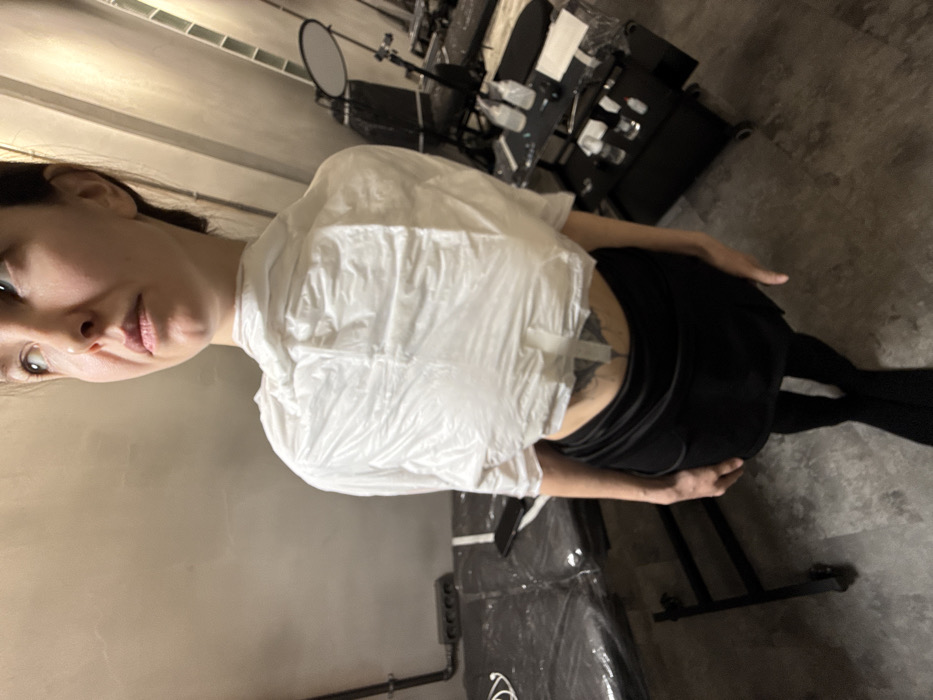 A female individual stands in a professional studio environment in Moscow, Russia. She is positioned slightly angled from the camera, looking upwards and to her right, with a neutral expression. Her upper torso is covered by a crinkled white plastic poncho or wrap, and she wears a black skirt over black tights. A fresh or recently completed dark-colored tattoo, resembling a symmetrical pattern, is visible on her lower back, partially covered by two horizontal strips of white medical tape.
To the left of the individual is a black examination or treatment table, draped with black plastic sheeting. A black power strip with three outlets is mounted on the light-colored wall above the table, with a white charging cable and a dark-colored smartphone resting on the covered surface. To the right, a multi-tiered black utility trolley with wheels holds various bottles and containers, also partially covered with plastic. Behind it, a black chair and a stand with a circular reflective disc are discernible, along with other indistinct dark equipment in the background. The studio features light-colored walls and a dark gray floor, illuminated by artificial light.