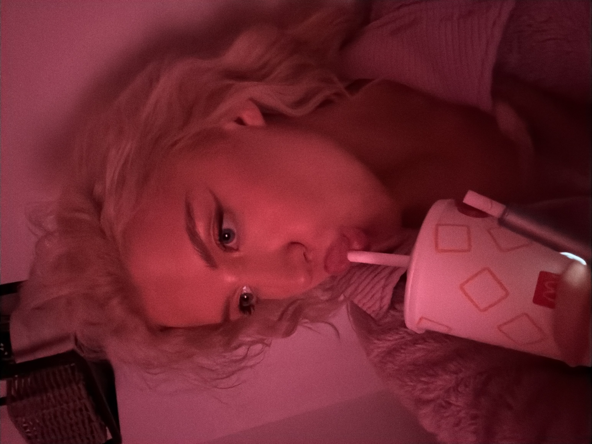 A person with blonde hair is shown from a low angle, holding a McDonald's cup with a straw in their mouth. The image is lit with a pink light. A black wicker basket is visible in the background. The city and country are not discernible from the image.