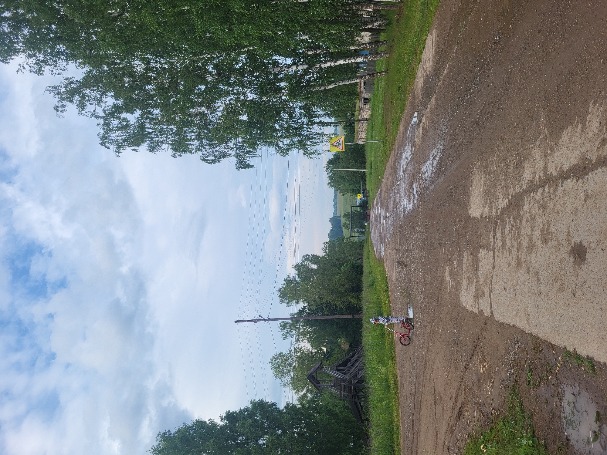 This media file depicts a rural scene in Zipunovo, Russia. A child, wearing a white and black patterned outfit and a helmet, is riding a small red bicycle on a wet, gravel road. The road is flanked by a grassy verge and tall trees with green foliage. In the background, there are more trees and a few buildings, suggesting a village or rural settlement. Power lines are visible stretching across the sky. There is a yellow road sign with a black symbol, possibly indicating a hazard or caution, on a pole to the left of the road. The sky is partly cloudy with patches of blue visible, and the overall lighting suggests it is daytime, likely in the afternoon. The presence of puddles on the road indicates recent rain. The scene evokes a sense of a peaceful, everyday moment in the countryside.