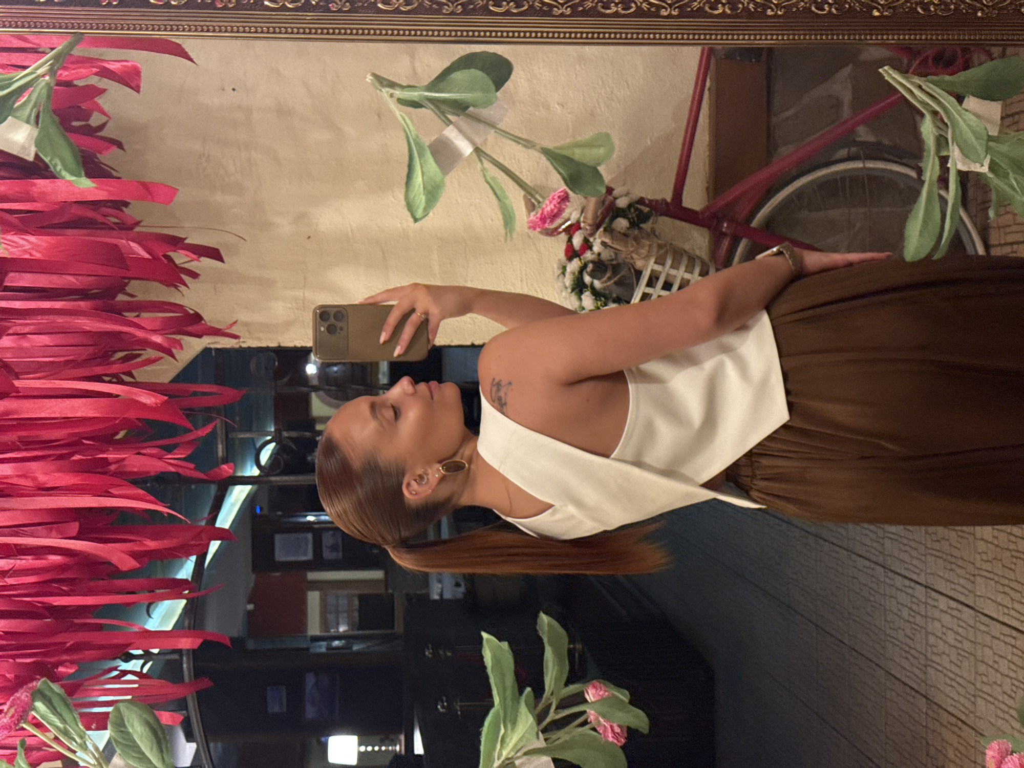 The image displays a mirror selfie of a young adult female taken inside what appears to be an indoor commercial establishment, possibly a restaurant or lounge. The woman, with reddish-brown hair neatly pulled into a sleek ponytail, stands with her back mostly to the viewer, turning her head slightly to her right (viewer's left) to reveal her profile. She wears a white sleeveless top with a distinctive criss-cross or wrap design on the back and loose-fitting, wide-leg brown trousers. Her left ear (as seen in the reflection) features multiple piercings, including a prominent oval gold-toned hoop earring. A small, dark tattoo is visible on her upper right arm/shoulder. She holds a dark-colored smartphone with a triple-lens camera in her left hand to capture the image. A thin ring is present on her left ring finger, and a metallic bracelet watch adorns her right wrist.
The selfie is taken in a large mirror bordered by an ornate gold-toned frame, which occupies the right side of the visual field. Several artificial green plant branches with pink flowers are visibly taped onto the surface of the mirror. Reflected in the mirror, behind the woman, is a red bicycle equipped with a white wicker basket filled with a bouquet of artificial red, white, and pink flowers. The wall reflected behind the bicycle appears to have a light, textured finish.
The upper portion of the scene features numerous red, possibly fabric, ribbons suspended from an unseen fixture, creating a dense overhead decoration. Some artificial green foliage is also visible among these ribbons on the far left. The background reflected on the left side of the mirror depicts a dimly lit interior with dark walls and furniture, including a table lamp with a glowing white shade on the far left and what appear to be framed pictures on a distant wall. The flooring throughout the visible area is a patterned surface, possibly parquet or decorative tile, with a geometric or interwoven design.