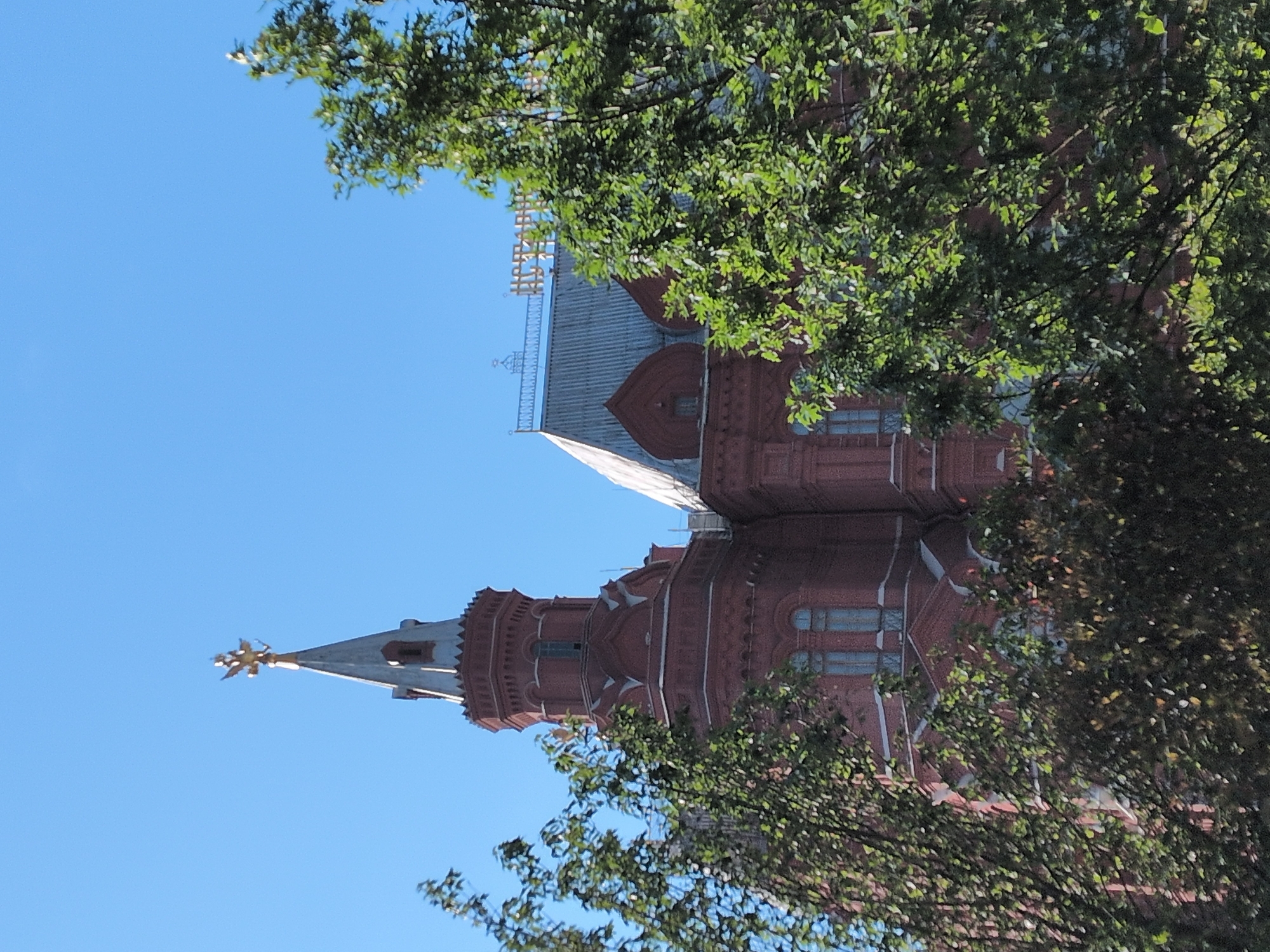 The image displays a grand, ornate red-brick building partially obscured by vibrant green tree foliage against a clear blue sky. The building features multiple towers and turrets, characteristic of a historical or monumental structure.
One prominent tower, situated on the left side of the frame (when viewed upright), culminates in a slender spire topped with a golden, star-like finial. The building's red brickwork exhibits intricate patterns and decorative elements, including arched and rectangular windows with reflective panes. The roof sections are grey, possibly metal, and are adorned with white, ornate detailing along their ridges and eaves.
Lush, green tree leaves and branches dominate the foreground and extend into the midground, casting dappled light and shadow and framing parts of the building. The bright, clear blue sky suggests a sunny day, likely morning or afternoon, given the even illumination and lack of harsh shadows. There are no people or discernible activities taking place. No visible text is present in the image.