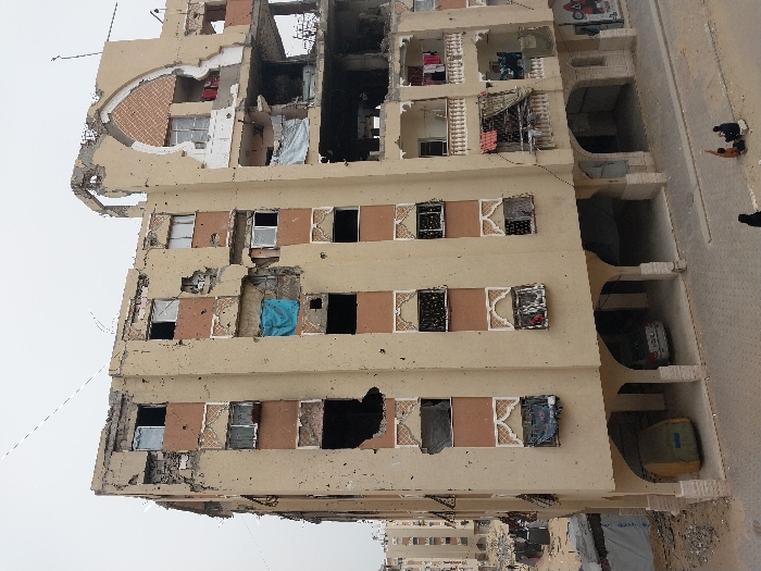 The image presents a severely damaged multi-story building in Khan Yunis, Palestinian Territory. The structure, painted primarily light brown with reddish-brown panels, shows numerous large apertures, missing wall sections, and widespread impact marks across its facade. Significant structural compromise is evident on the upper right, where a large decorative arch is partially collapsed, exposing interior rooms and structural elements. Multiple windows are shattered or missing panes; some are covered with assorted fabrics, notably a blue tarp on the third floor and white sheets on balconies on the right side. Several balconies with white balustrades are present on the right, displaying hanging laundry. The ground floor features open arched bays, accommodating a silver sedan and a covered object under a green-yellow tarp. A paved street occupies the foreground, where three individuals are seated on the lower right: one in dark clothing and two in lighter garments, appearing to interact or rest. In the background to the left, further damaged buildings and debris piles are visible, with several distant human figures moving amongst them. The sky is uniformly overcast.