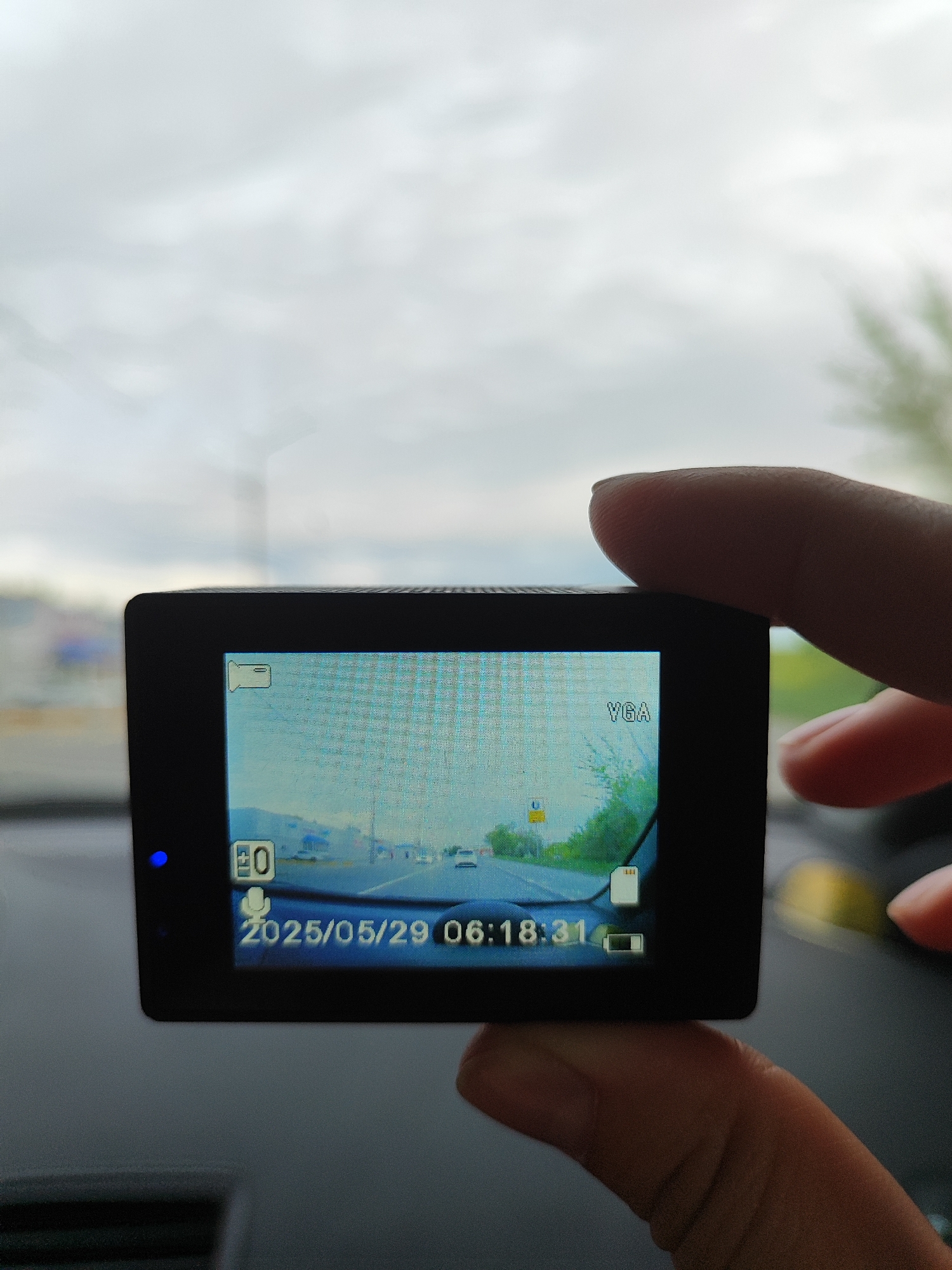 A black dashboard camera is held in a person's hand. The camera displays a recording of a roadway taken from inside a vehicle, dated 2025/05/29 at 06:18:31. The sky is cloudy. The location is Unknown, Unknown.