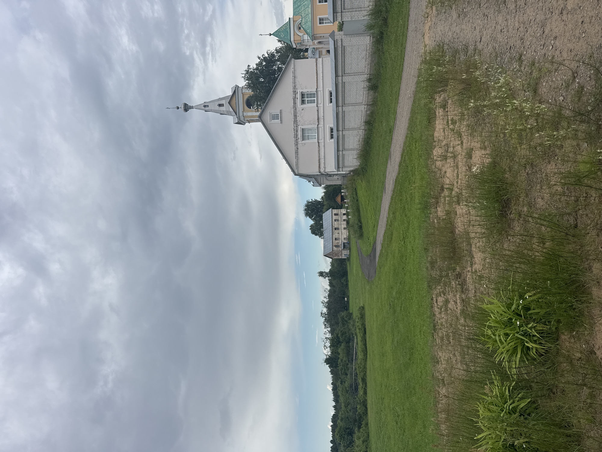 The image captures a serene, slightly overcast day in the village of Рассвет, Russia. In the foreground, a dirt path winds through a grassy area with tall weeds and some leafy green plants. The path leads towards a cluster of buildings, dominated by a light pink, two-story structure with a distinctive white brick wall in front of it. To the right of this building, a white and yellow bell tower, topped with a spire and a small cross, rises against the cloudy sky. Adjacent to it, another building with a green roof is partially visible. Further in the background, a line of trees creates a dark green backdrop, and to the left, a low bridge spans a body of water surrounded by vegetation. The overall scene suggests a quiet, rural atmosphere with traditional architecture.