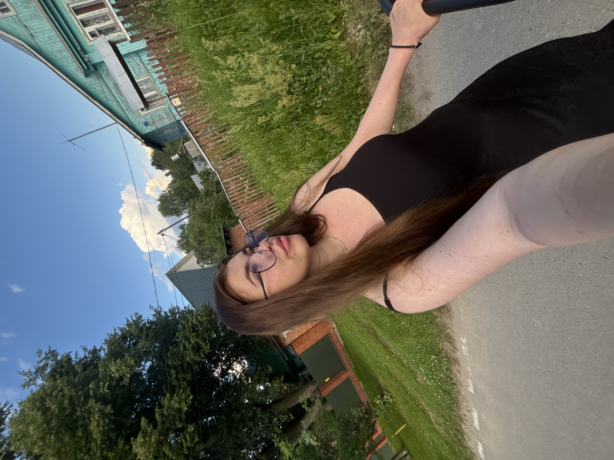 A young woman with long brown hair and glasses is shown from the chest up, looking to her right. She is wearing a black dress and is standing outside. Behind her, there is a street, a wooden fence, green grass and trees, and a wooden house painted turquoise. The sky is blue with white clouds. The scene appears to be in a village or rural area, possibly in the city of Мещёры, Russia, given the presence of a traditional wooden house and the general environment.