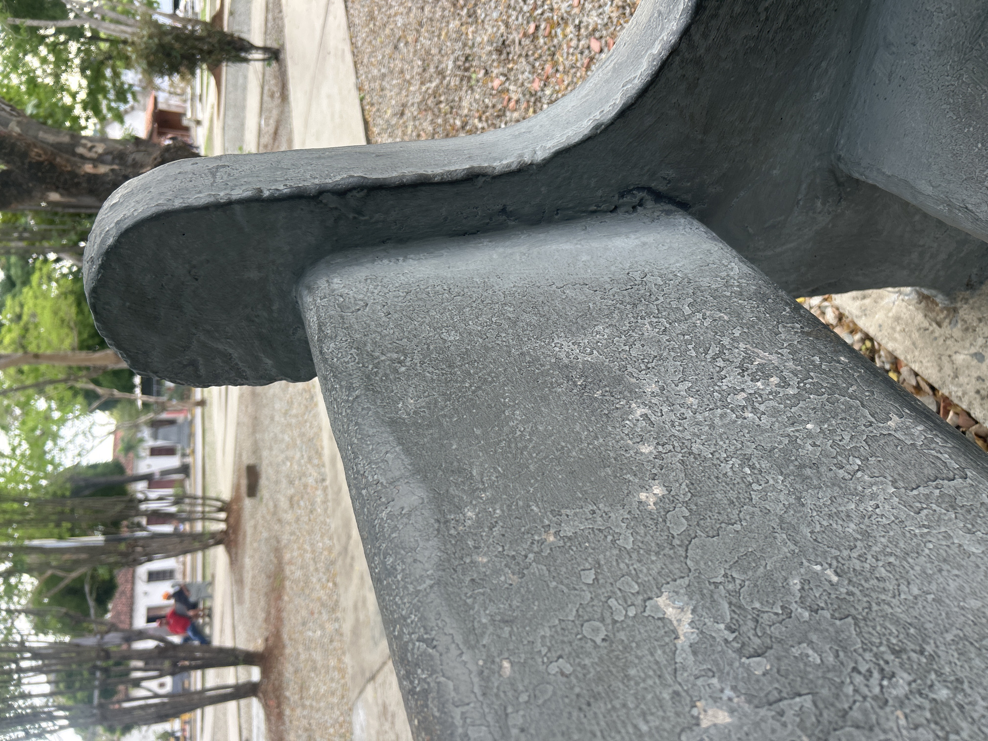 The image shows a close-up of a weathered, grey, concrete bench in a park. The bench has a rough texture with areas where the paint or coating is peeling. In the background, a park setting is visible with trees, paved walkways, and buildings with red-tiled roofs, characteristic of Santa Ana, Venezuela. Two individuals are seated on another bench in the distance. The ground is covered with small stones and fallen leaves. The focus is on the foreground bench, highlighting its texture and material.