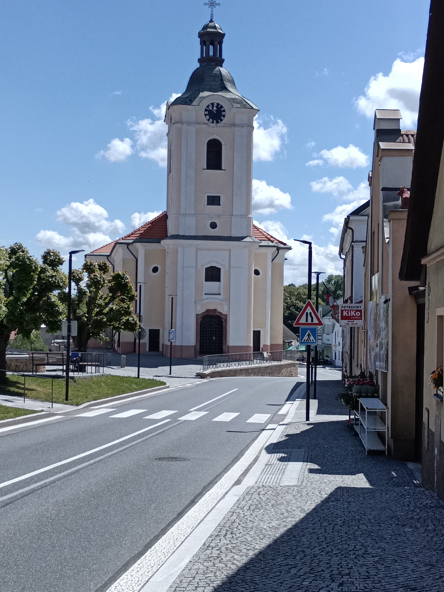 The image depicts a street scene in Dalovy, Czechia, on a clear, sunny day with a blue sky and scattered white clouds. A prominent church with a light beige facade and white trim dominates the center, featuring a tall square bell tower topped with a dark, possibly copper or slate, conical roof culminating in an orb and a cross. A clock face on the tower shows approximately 11:15 or 11:20. The church's main body has a red-tiled roof and arched windows.
In the foreground, an asphalt road with a double solid white line runs towards the church, featuring a pedestrian crosswalk (zebra stripes). To the left of the road, there is a grassy area with several mature, heavily pruned trees, a park bench, and modern black streetlights. A low stone wall forms the boundary of this area.
On the right side of the road, a cobblestone sidewalk extends along a row of multi-story buildings. Deep shadows are cast across a significant portion of the sidewalk. One building features a red and white sign for "POTRAVINY Kiko EXPRES" with listed hours "DENNĚ 7:00 - 20:30." Below this sign, a white metal shelving unit is placed on the sidewalk. Window boxes with flowers adorn the windows of these buildings. A red, possibly ceramic, cow figurine is visible on the upper part of one building's facade or roofline. Two traffic signs are mounted on a pole near the right curb: a triangular red-bordered sign indicating a narrowing road and a square blue sign denoting a pedestrian crossing. No people are visible in the scene.