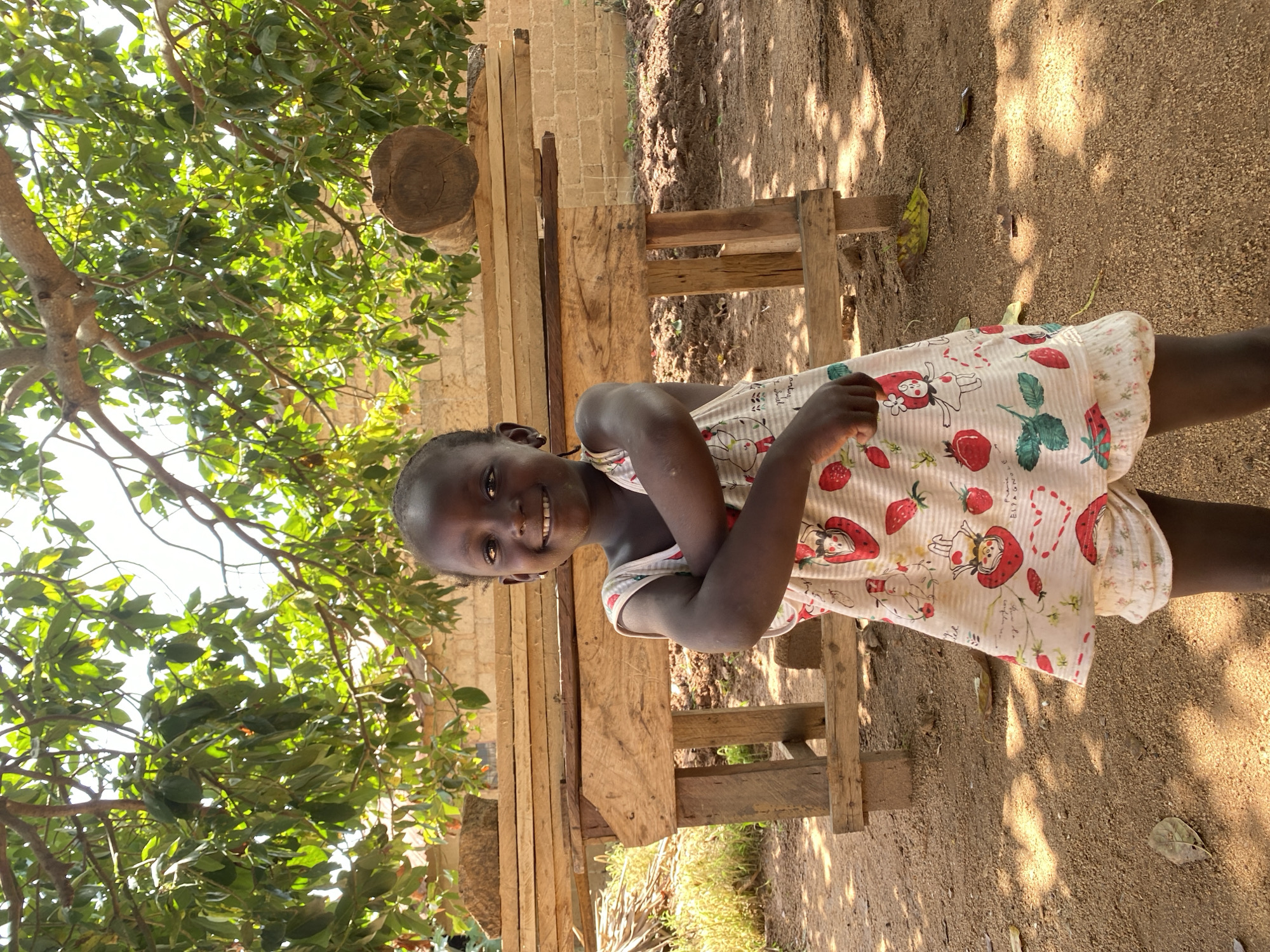 A young girl stands outdoors, smiling. She wears a light-colored dress patterned with strawberries and cartoon figures. She is positioned in front of a roughly constructed wooden bench or structure. A tree with dense green foliage partially obscures the view above and to the left. The ground is dirt. A brick structure is faintly visible behind the wooden structure. The photograph was taken in Riyom, Nigeria.