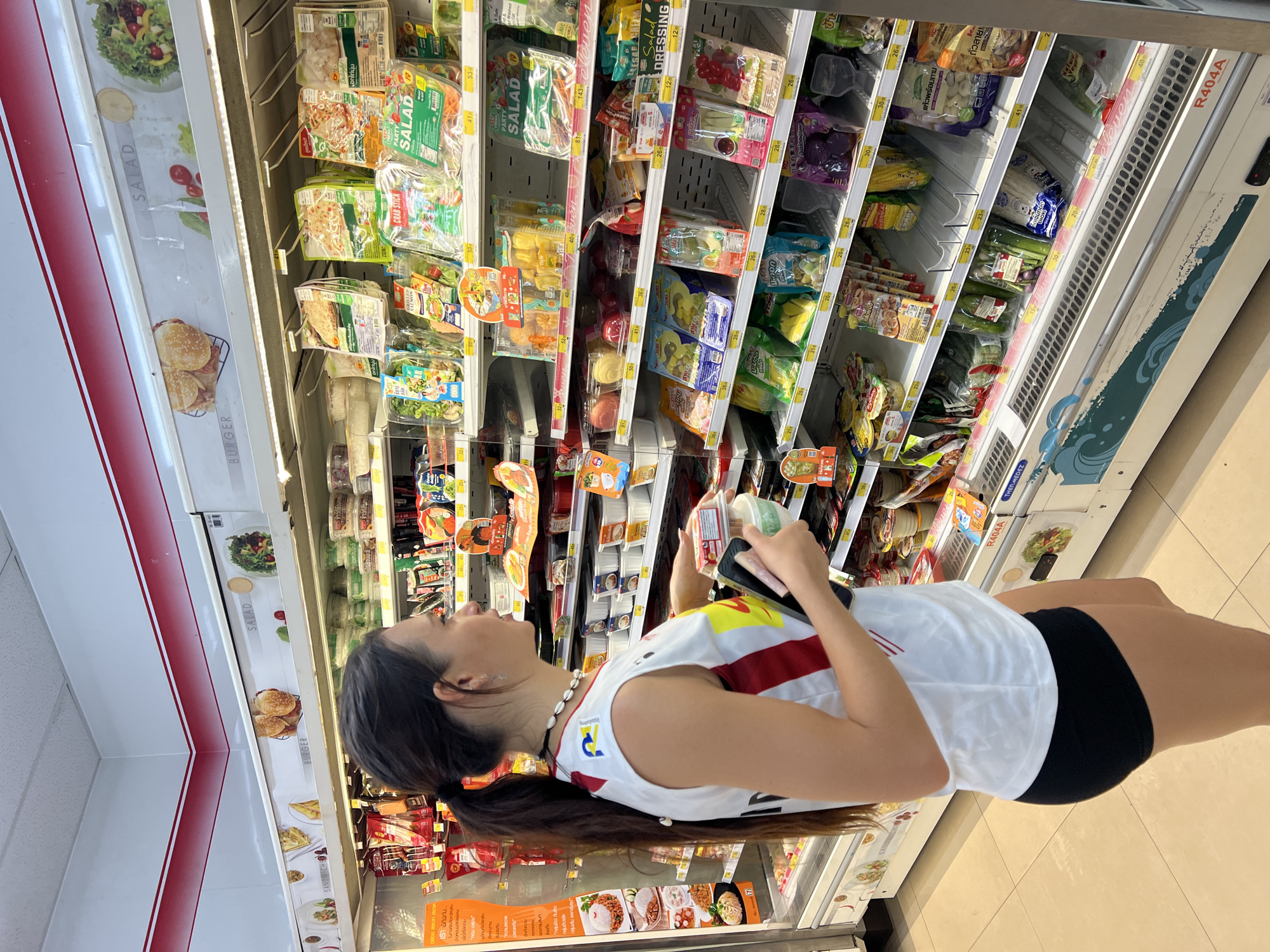 A woman stands in front of a refrigerated display case in a convenience store in บ้านบางรักษ์, Thailand. She wears a white t-shirt with red stripes, black shorts, and a shell necklace. Her hair is pulled back in a ponytail. She holds a container of food in her hands, looking at the contents of the display case. The display case contains a variety of pre-packaged salads, entrees, and ingredients. The case is labeled with prices in Thai currency. Burgers and salads are pictured on the door of the fridge. The floor is tiled.