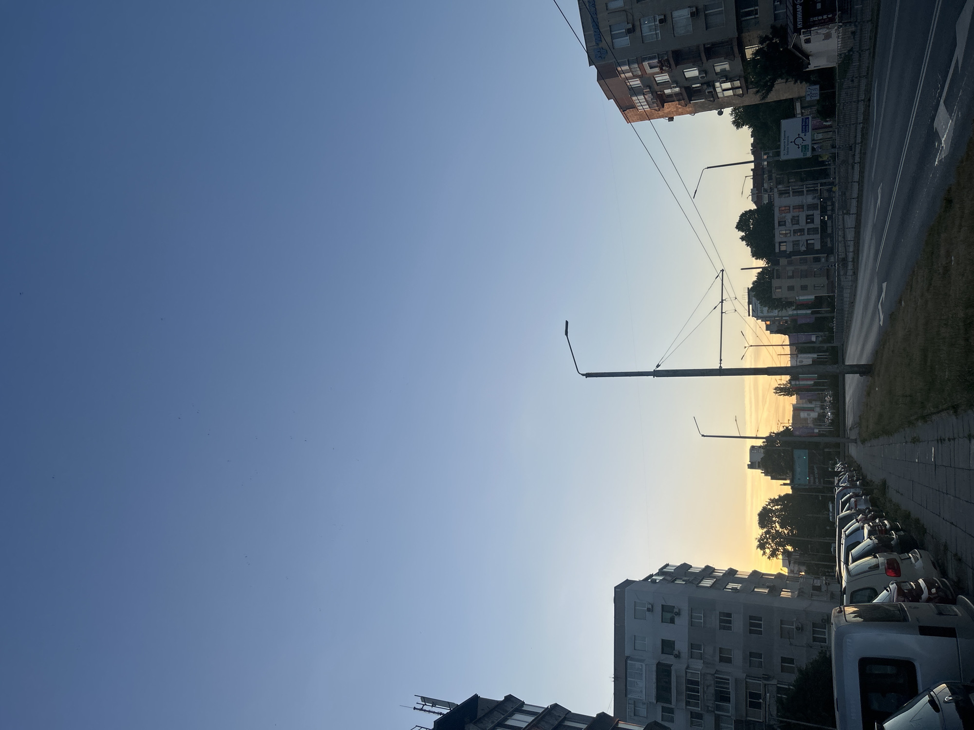 The image depicts a street scene in Varna, Bulgaria, during what appears to be either sunrise or sunset, indicated by the warm glow on the horizon. The sky is a clear blue, transitioning to soft oranges and yellows near the buildings.
On the left side of the street, a multi-story residential building with numerous windows and balconies is visible. In front of this building, a row of parked cars is lined up along a sidewalk. The cars are of various makes and models, predominantly sedans and vans.
Across the street, on the right, is another multi-story building, also with many windows and visible air conditioning units. A street sign with Cyrillic text is present, indicating directions to locations such as "Ulp. Vl. Varnenchik" and "Knyaz Boris I." This suggests the location is within or near the city of Varna.
The street itself has multiple lanes of asphalt, with directional arrows painted on the surface. Overhead, streetlights and the wires of a tram or trolleybus system are visible, suggesting it's a public transportation route. Trees line the street intermittently, with some taller trees in the background.
There are no people clearly visible in the frame. The scene is static, capturing a moment of quiet urban activity. The overall impression is one of a typical residential and commercial street in a Bulgarian city, bathed in the soft light of dawn or dusk.