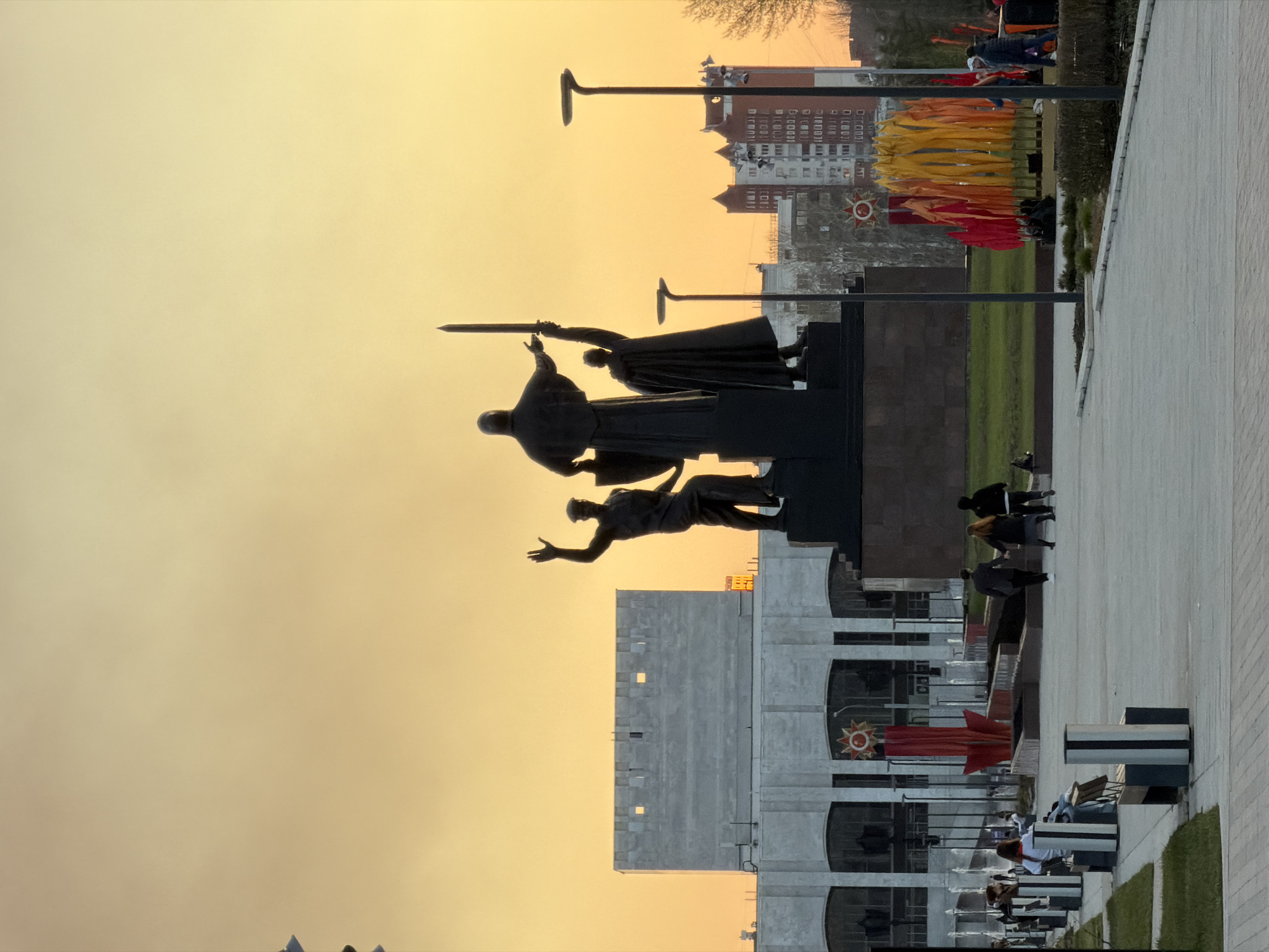 The image shows a view of the Perm Victory Monument in Perm, Russia, at dusk. A bronze statue of three figures - a kneeling soldier, a standing man with a sword, and a woman - stands prominently against a hazy yellow sky. In the background, there is a multi-story building and other structures. Below the statue, there's a grey, modern building with a red star emblem. People are walking on a paved area with benches, fountains and trimmed hedges. A row of flag banners decorated with red and yellow colors is visible in the background. Lampposts rise above the scene.