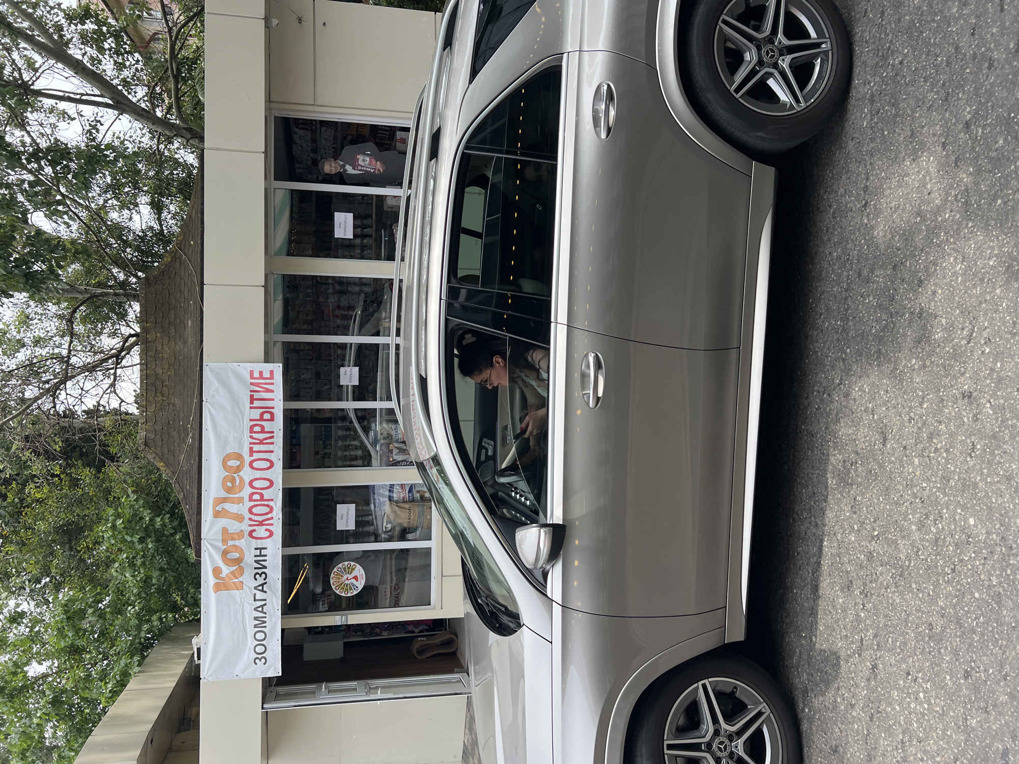 A woman is driving a silver SUV in Sochi, Russia. She is wearing glasses and has her hair pulled back. The SUV is parked on a street in front of a building with a banner that reads "Кот Лео" (Kot Leo) and "ЗОOMAGAZIN СКОРО ОТКРЫТИЕ" (Zoo store opening soon). There is a cardboard cutout of a man in a suit holding a book in the window of the building. The building appears to be a shop, possibly a pet store given the "zoo store" text. The street is paved and there are trees and buildings in the background.