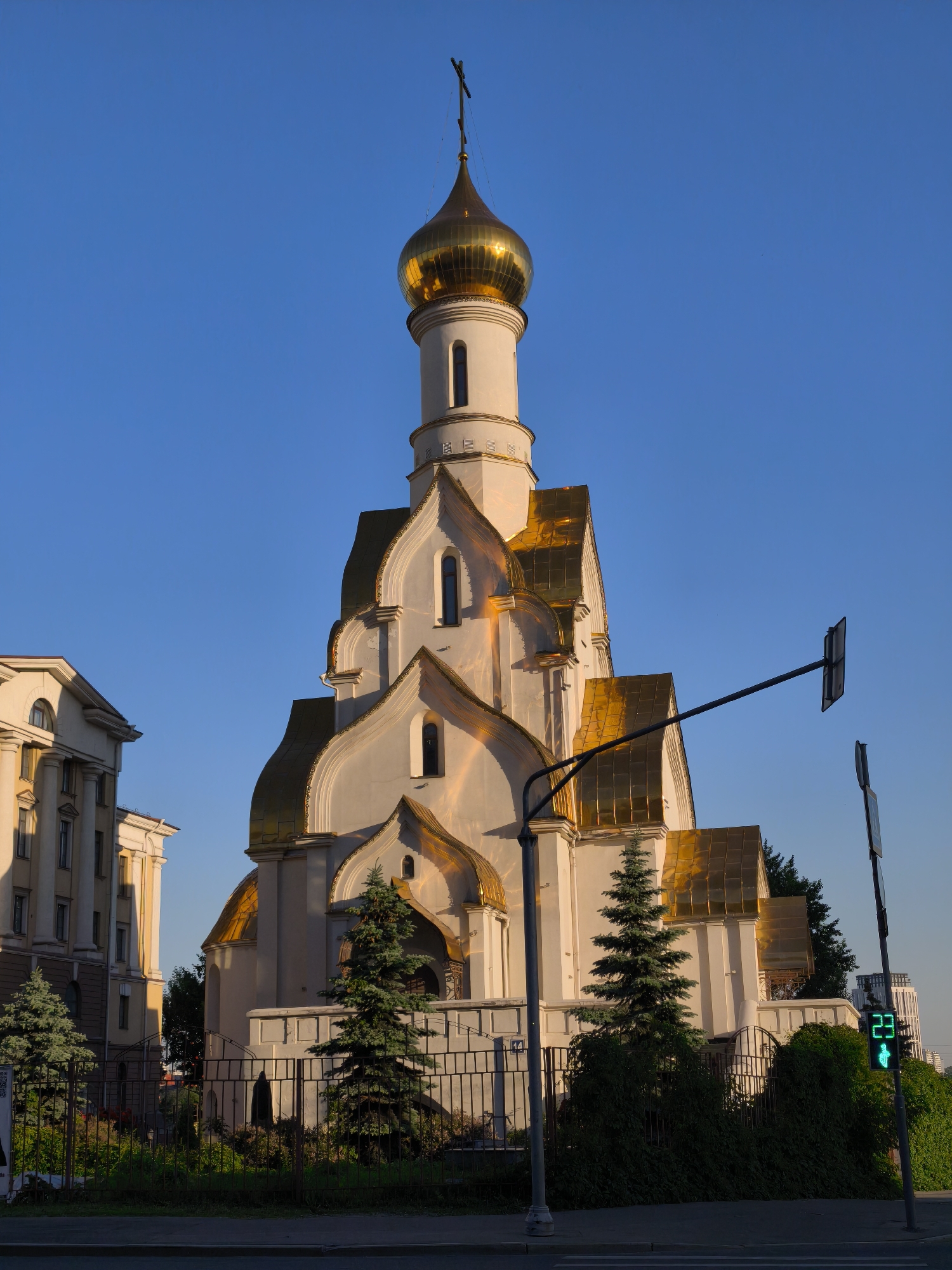 This image depicts the Church of the Holy Blessed Prince Alexander Nevsky, situated at the Moscow State Institute of International Relations (MGIMO) at Prospect Vernadskogo, 76, Moscow, Russia. The church, a prominent white edifice with traditional Russian Orthodox architectural elements, is bathed in warm, direct sunlight under a clear, cloudless blue sky, indicating either early morning or late afternoon.
The central feature is a tall, slender tower culminating in a brightly gleaming golden onion dome topped with a dark Orthodox cross. Below the main dome, multiple tiers of golden, curved roofs create a distinctive wave-like or stacked appearance, contrasting with the smooth, light-colored (likely white or very light beige) walls of the church. The church walls feature tall, narrow, arched windows.
To the left of the church, a large, multi-story building with classical architectural details, including pilasters and arched window openings, is partially visible. This building is also light-colored and appears to be part of an institutional or academic complex, consistent with the MGIMO campus.
In the foreground, a dark asphalt road and a paved sidewalk are visible. A black metal fence delineates the church grounds from the public thoroughfare. Within the fenced area, several mature, dark green coniferous trees (likely spruces) are planted, alongside lower bushes and groundcover.
On the right side of the image, a dark pole supports a pedestrian traffic light displaying a green "walk" symbol with the number "23" in green digits, indicating the seconds remaining to cross. Another, smaller sign pole stands nearby. Extending from the left side over the road is a long, dark, horizontal boom-like structure with a rectangular panel, possibly a gantry for traffic signals or surveillance equipment. In the distant background on the far right, a modern, light-colored high-rise building is faintly discernible. No people are present in the scene.