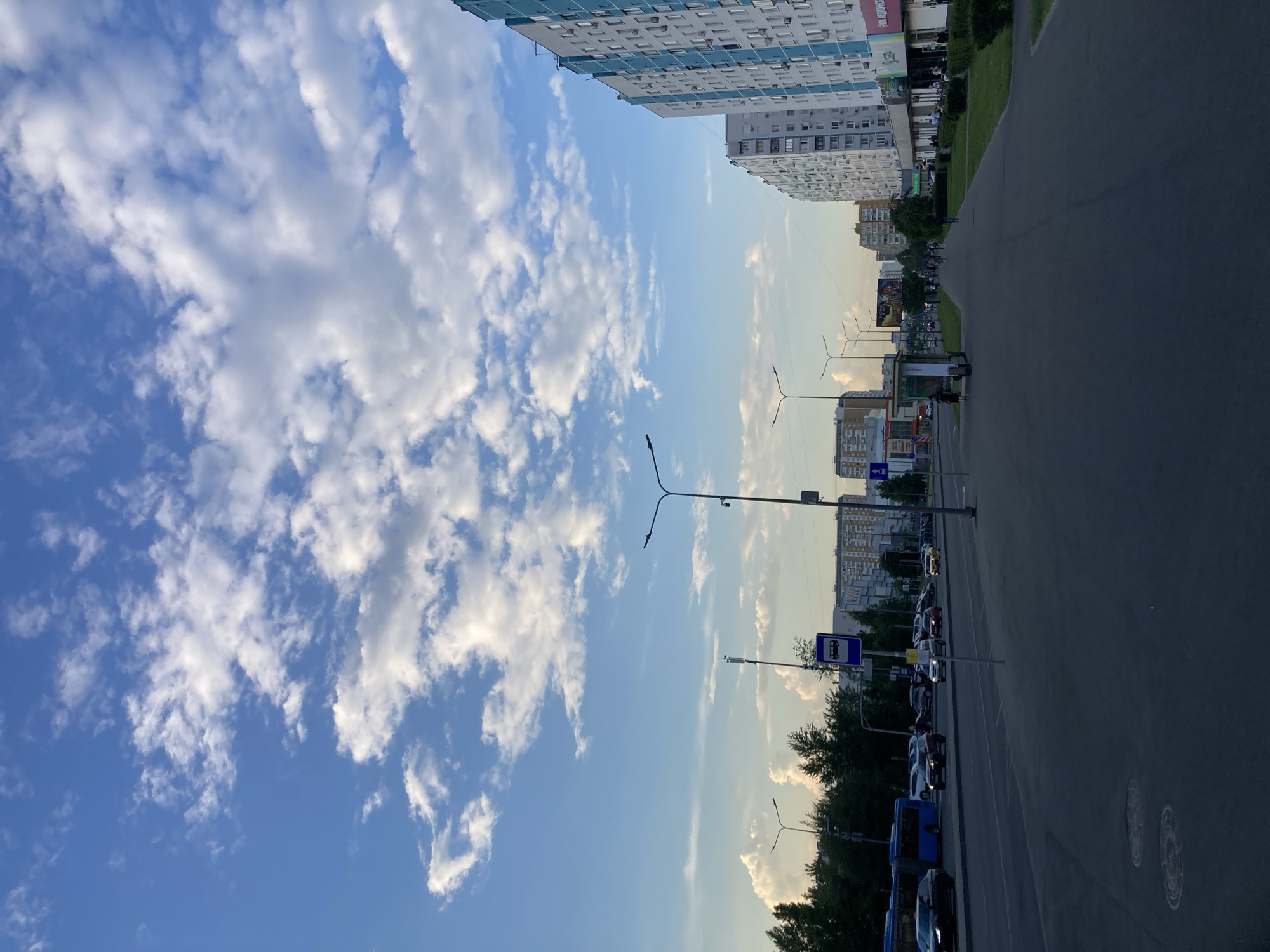 This image depicts a wide street in Moscow, Russia, under a bright blue sky with scattered white clouds. On the right side of the street are several tall, multi-story apartment buildings, some with light-colored facades and others with a distinctive light blue and white design. The buildings have numerous windows and air conditioning units. A sidewalk runs along the front of these buildings, lined with neatly trimmed green bushes. Several shops and businesses are visible at the ground level of these buildings.
On the left side of the street, there is a multi-lane road with moderate traffic. Cars, including a blue bus, are driving or parked. A bus stop sign is visible next to the road. Beyond the road, a line of trees forms a green border. In the distance, more apartment buildings and some billboards can be seen. The overall impression is that of a typical urban residential and commercial area. The lighting suggests it is daytime, likely late afternoon or early evening due to the angle of the sun and the quality of the light. There are a few pedestrians visible on the sidewalk and near the shops.