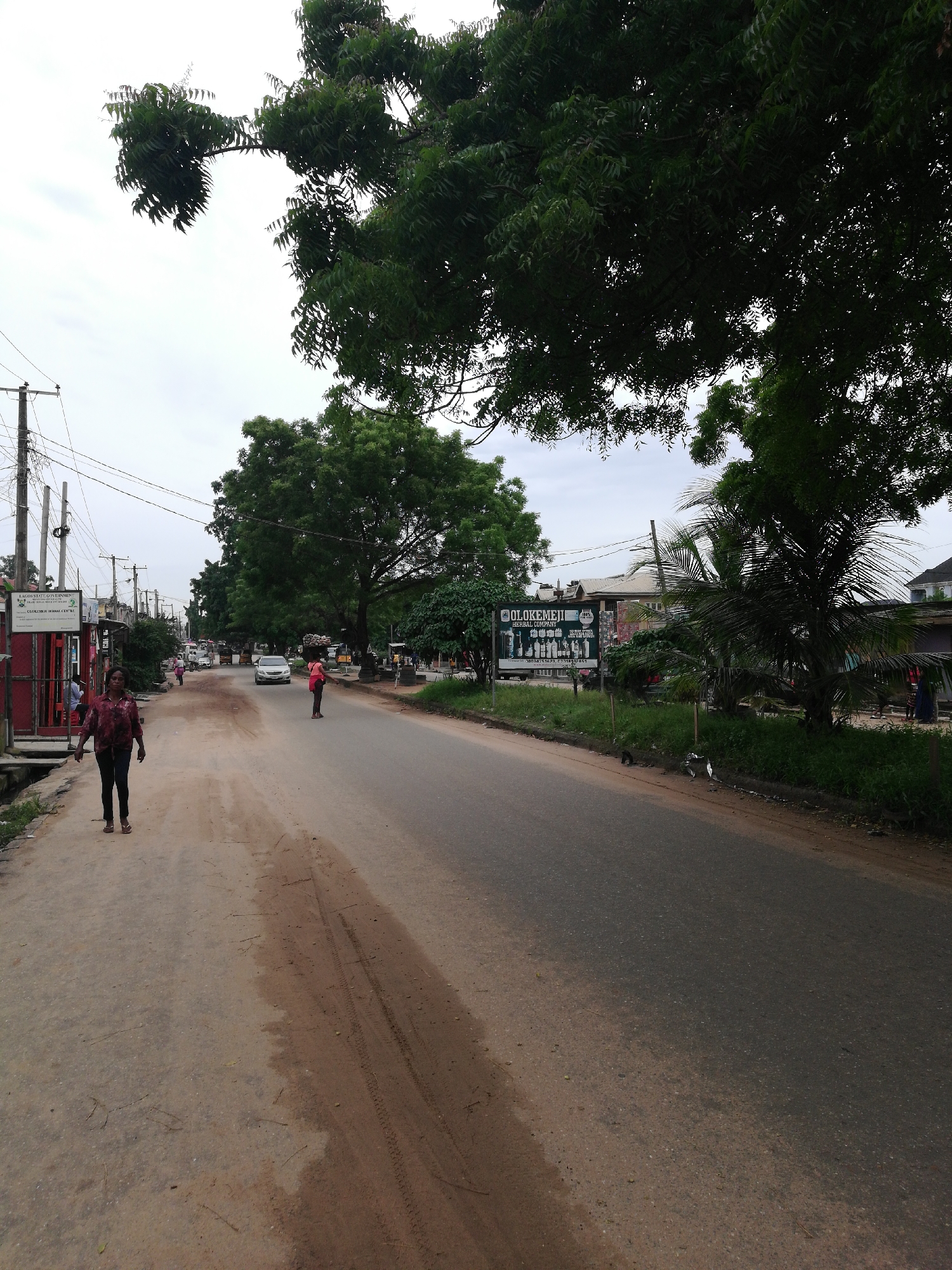 The image depicts a street scene in Lagos, Nigeria. The road is paved but appears worn with patches of dirt and tire tracks along the shoulder. A woman walks on the left side of the road, while another person stands in the middle distance wearing an orange top. A car is visible further down the road. A large billboard advertising "Olokemeji Natural Company" is on the right side of the street, bordered by green vegetation. Trees overhang the road, and power lines are visible above. Buildings are seen in the background, suggesting an urban or semi-urban environment. The sky is overcast.