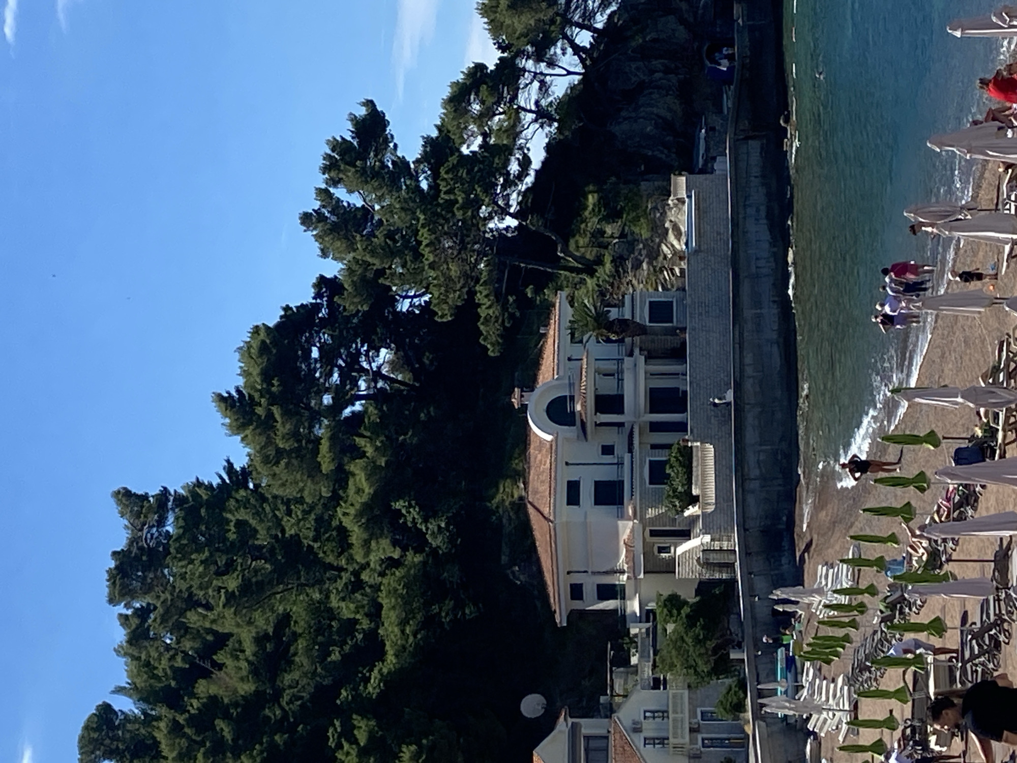 The image depicts a sunny day at a beach in Petrovac na Moru, Montenegro, with a prominent white villa situated on a hillside overlooking the water. The villa features a red-tiled roof, balconies, and large dark-shuttered windows, with a satellite dish visible on an adjacent building. Lush green trees, characteristic of the Mediterranean coast, cover the hillside behind the villa.
The beach itself is lined with rows of empty lounge chairs and closed umbrellas, suggesting a resort setting. A few people are visible on the sand, some walking towards the clear turquoise water, while others are seated on chairs or near the shoreline. The water is calm, with gentle waves lapping the shore. In the distance, a rocky outcrop protrudes into the sea, further enhancing the scenic coastal landscape. The sky is a clear, vibrant blue, indicative of fair weather.