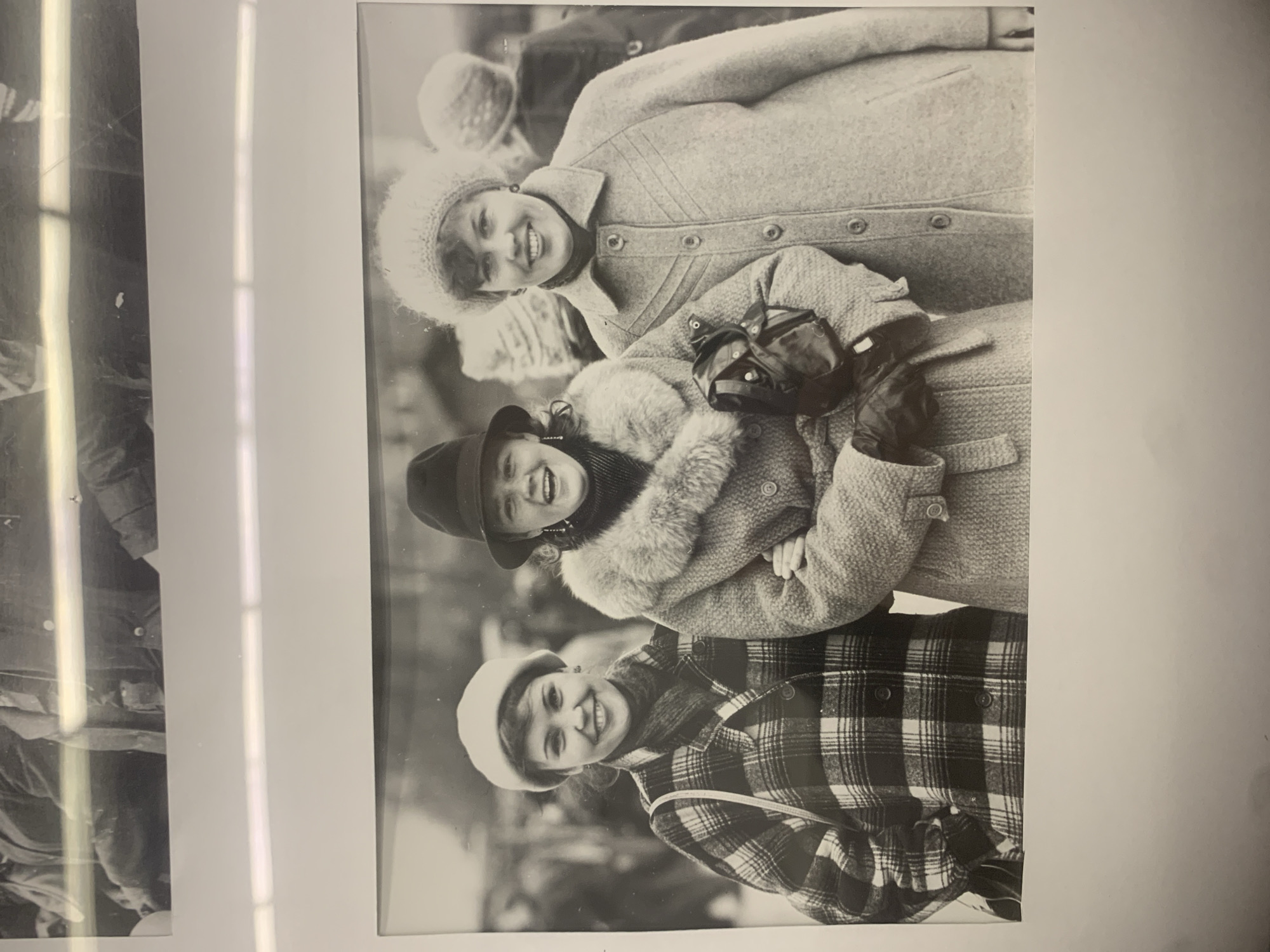A black and white photograph depicts three women bundled in winter clothing, likely taken outdoors. The topmost woman wears a light-colored knit hat and a textured coat with large buttons. The middle woman wears a hat, a fur-collared coat, and leather gloves. The bottom woman is dressed in a plaid coat and a hat. All three are smiling at the camera. The setting suggests a winter environment with snow or a similar backdrop. There is no information to indicate it's in Izhevsk, Russia.