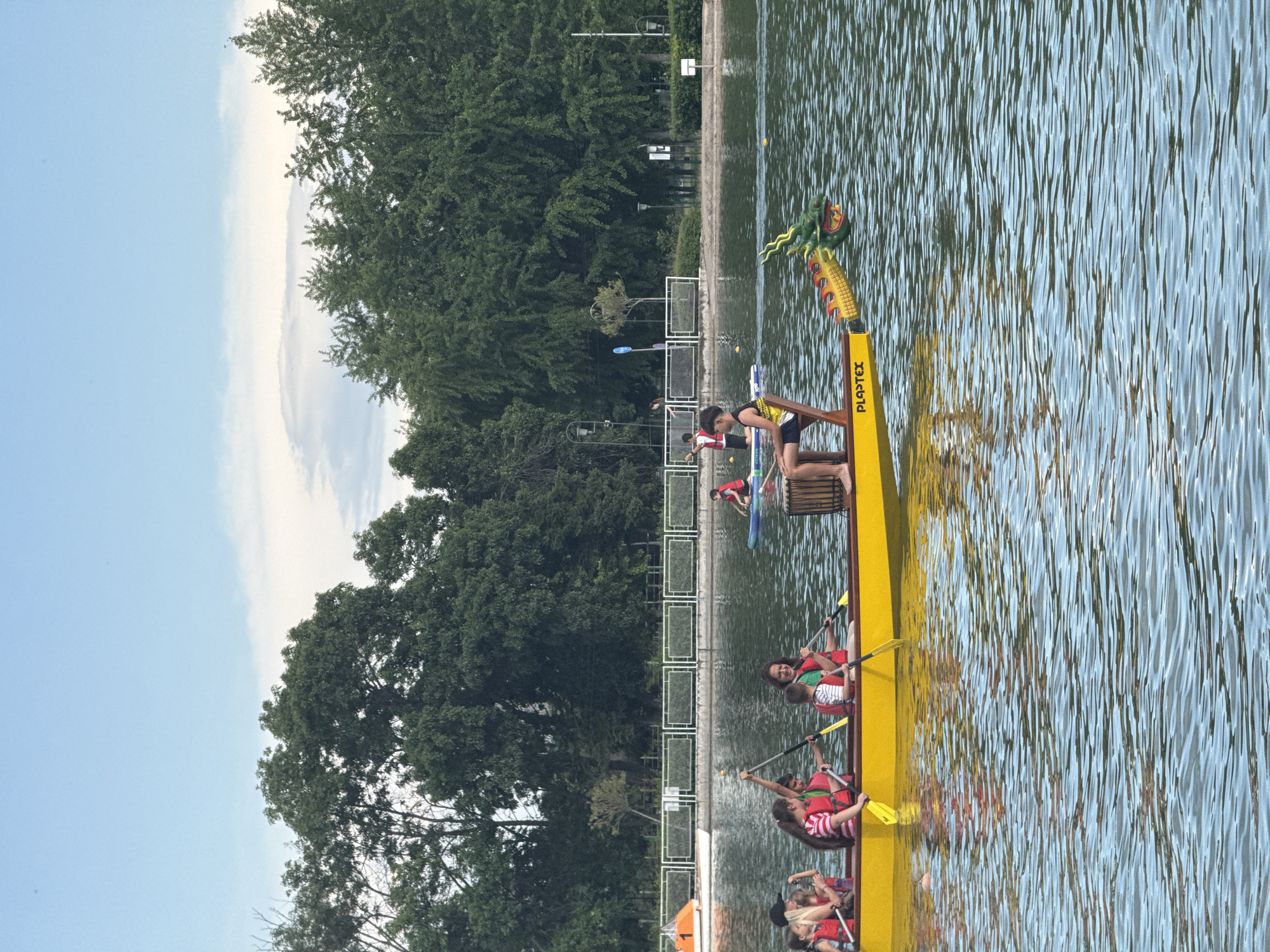 A horizontal medium shot captures a bright yellow dragon boat on the Plovdiv Rowing Canal in Bulgaria. The boat is filled with individuals wearing life vests and holding paddles, suggesting a rowing activity. At the front of the boat, a person stands, possibly navigating or directing. The boat features a decorative dragon head at the bow. The water is textured with ripples. The canal is bordered by a grassy bank with trees and a metal fence. The sky is blue with some clouds.
