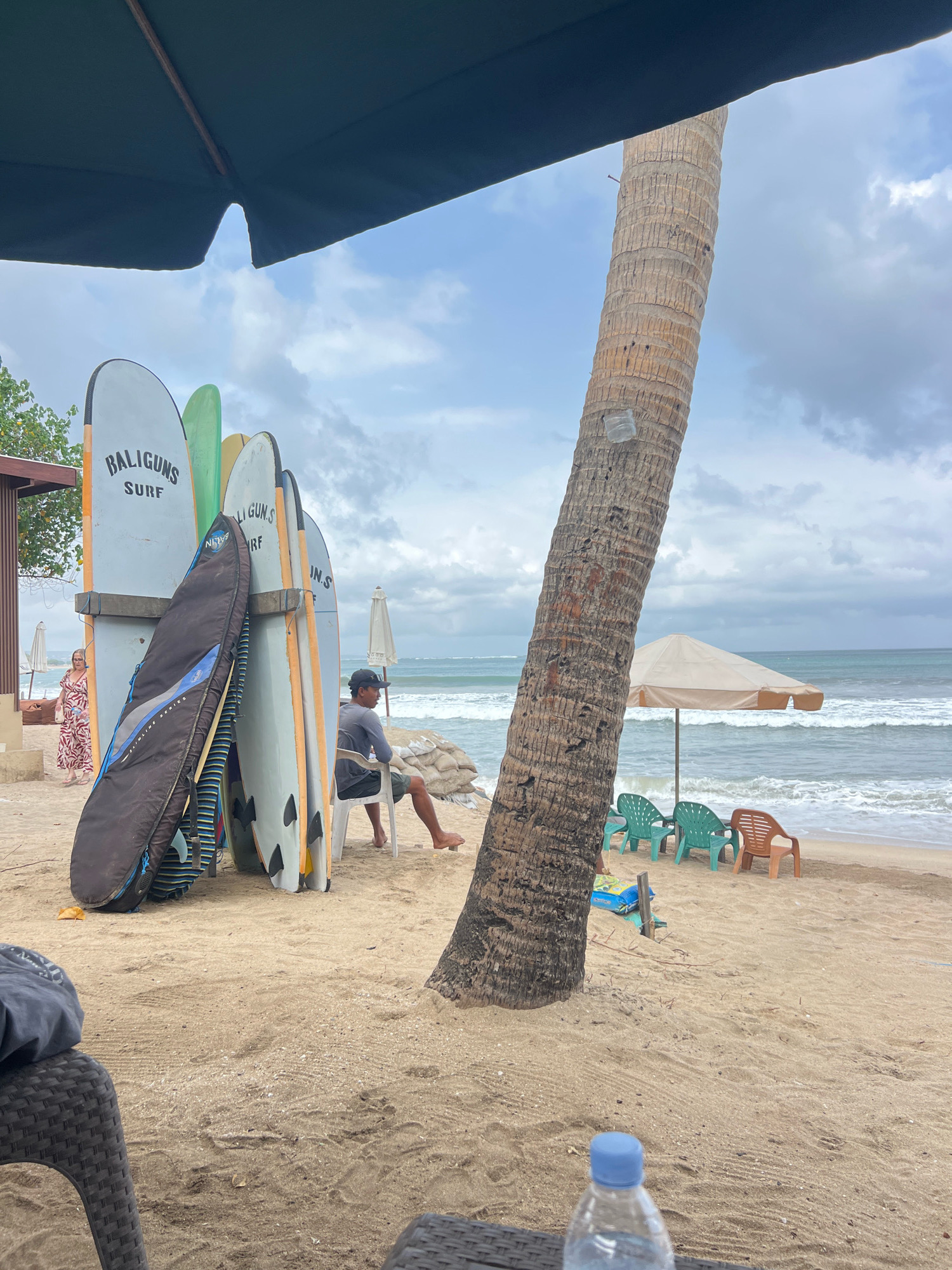 This is a medium shot of a sandy beach in Kuta, Indonesia, with several surfboards leaning against a wooden rack near a palm tree. In the foreground, a dark woven chair and a partially visible table are in front of a plastic water bottle. To the left of the surfboards, a woman in a colorful dress walks away from the camera. To the right of the surfboards, a man wearing a grey t-shirt and shorts sits on a white plastic chair facing the ocean, with sandbags piled behind him. In the mid-ground, several plastic chairs and a beige beach umbrella are set up on the sand facing the wavy ocean. The sky is overcast with scattered clouds.