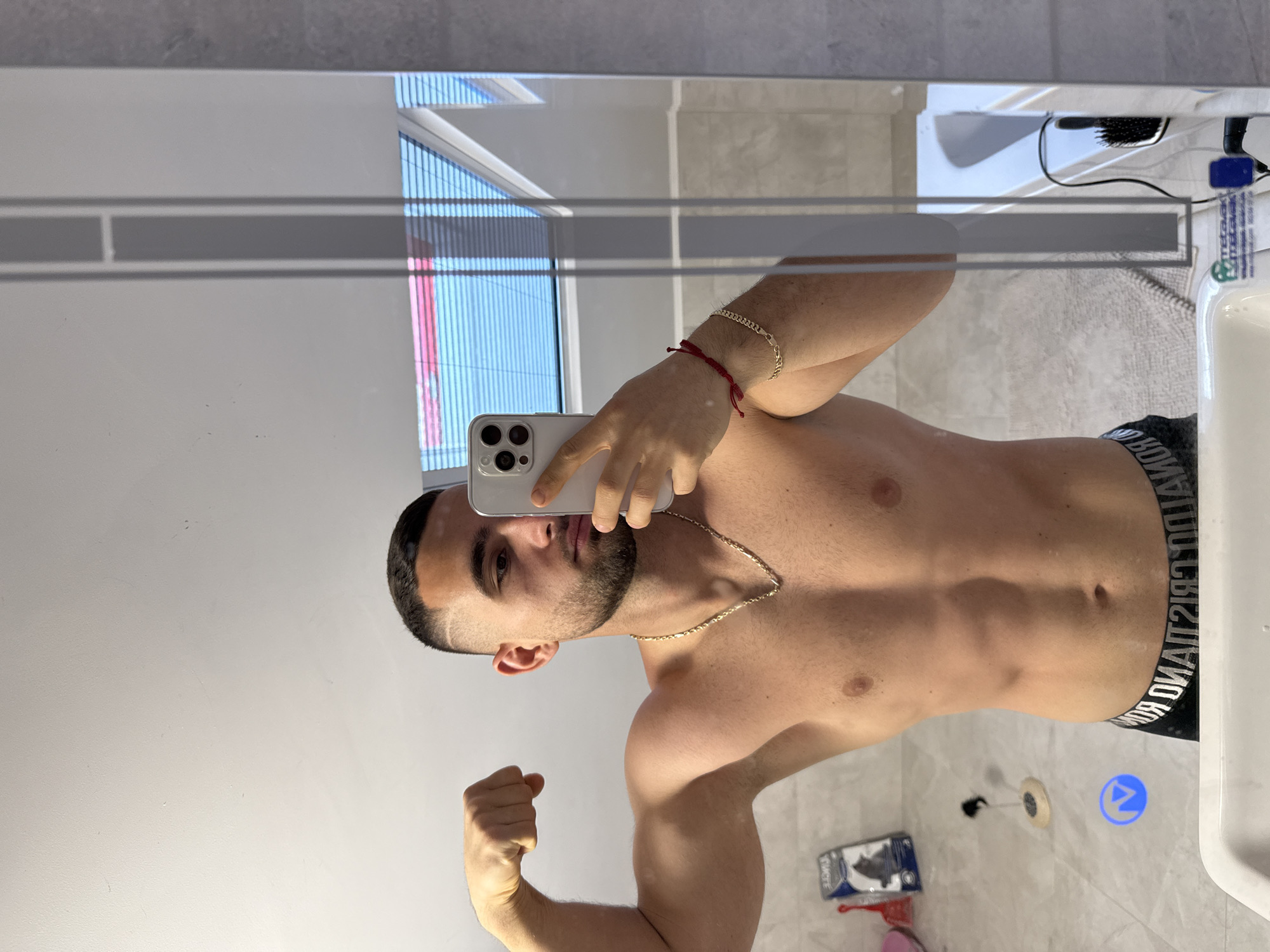 A shirtless man with a short haircut and beard is taking a mirror selfie in a bathroom. He is holding a smartphone with multiple cameras. He is wearing a gold necklace and bracelets. The bathroom has gray tiles, a sink, and a window with blinds. The location in Gabrovo, Bulgaria, is unknown.