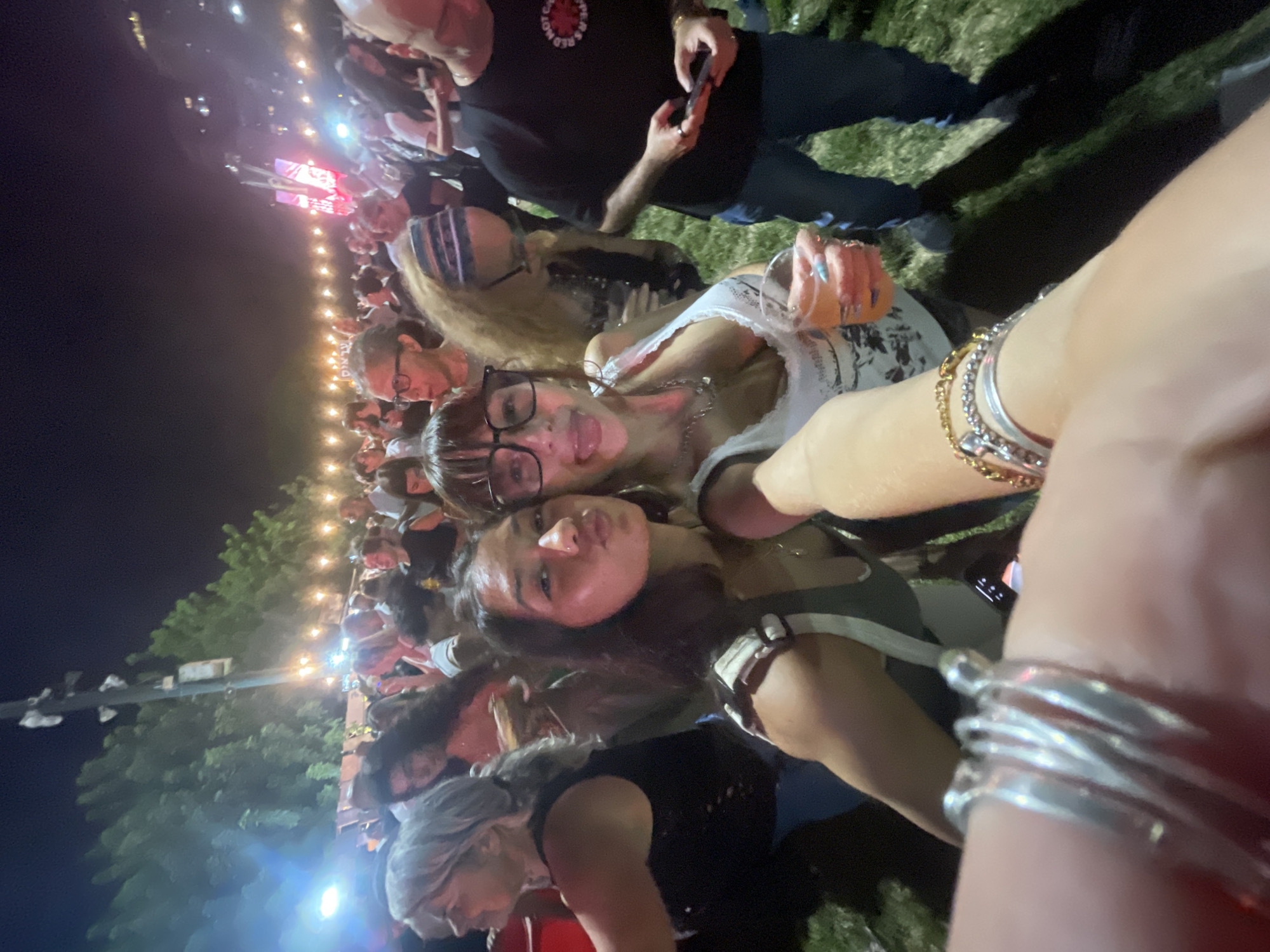 In an outdoor nighttime setting, possibly a festival or concert in Petah Tikva, Israel, two young women are taking a selfie. The woman on the left is wearing a green tank top and has her arm outstretched to capture the photo. She is making a puckering gesture with her lips. The woman on the right is wearing a white lace-trimmed tank top and large-rimmed glasses. She has her tongue sticking out and is holding a drink in her right hand. Both women are adorned with bracelets and rings. The background is filled with a crowd of people, some of whom are illuminated by string lights and spotlights. Trees are visible in the distance, and the ground appears to be covered in grass. The overall atmosphere suggests a lively social gathering.